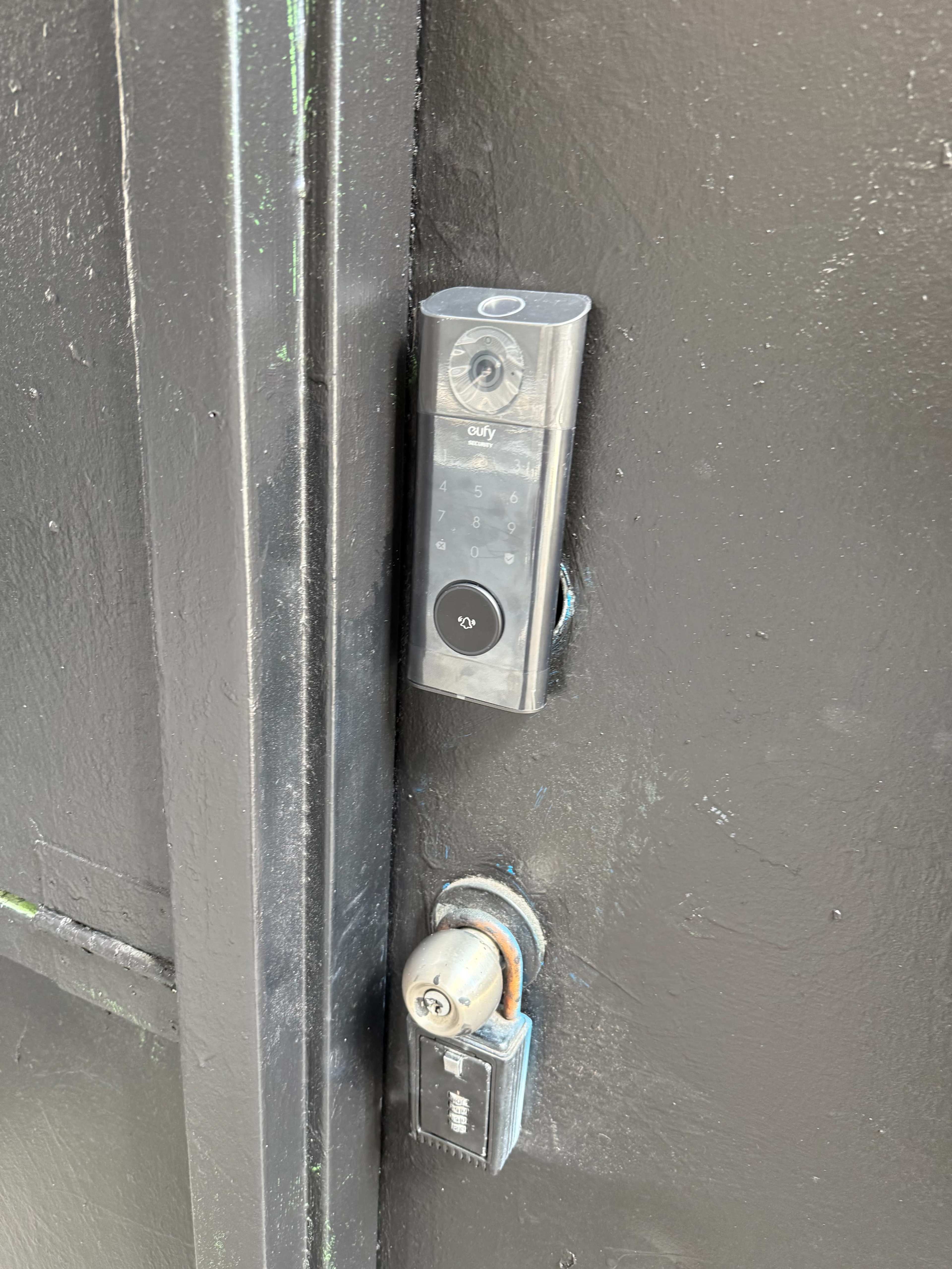 The image shows a smart door lock with a numeric keypad installed on a black door, alongside a traditional key lock.