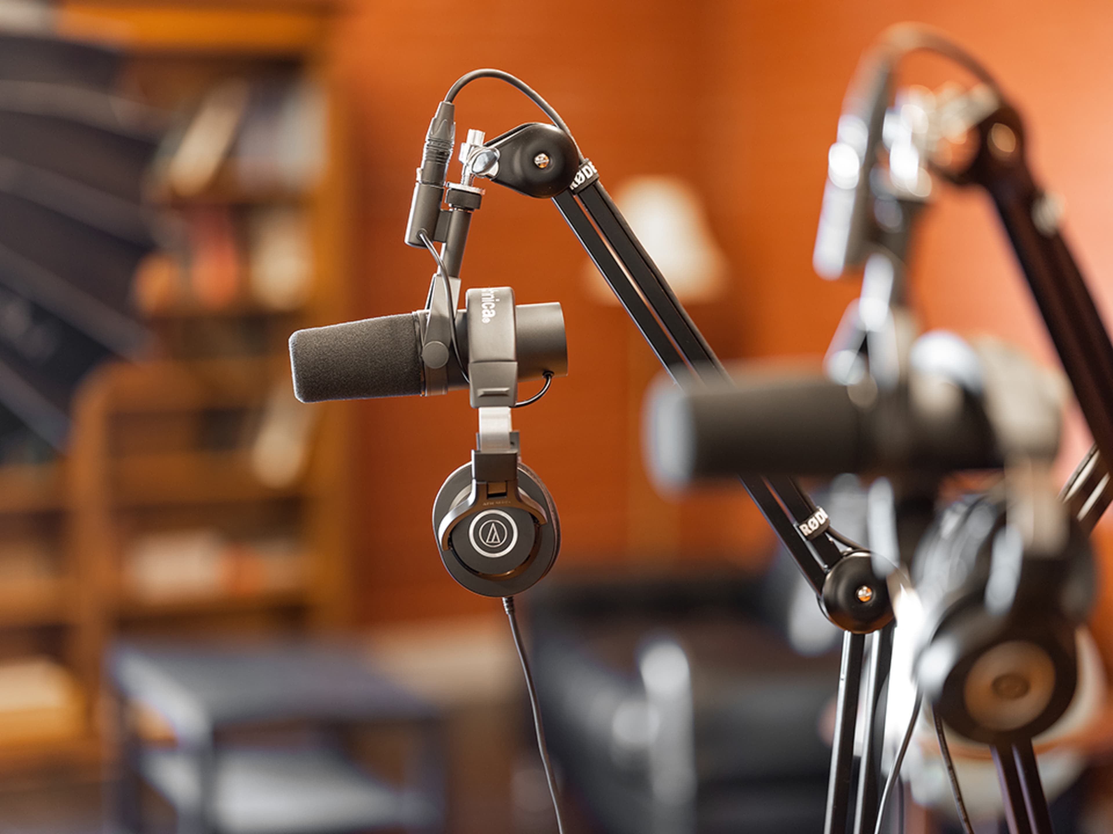 A close-up of a microphone and headphones mounted on adjustable microphone stands, with a blurred background of furniture and shelves.