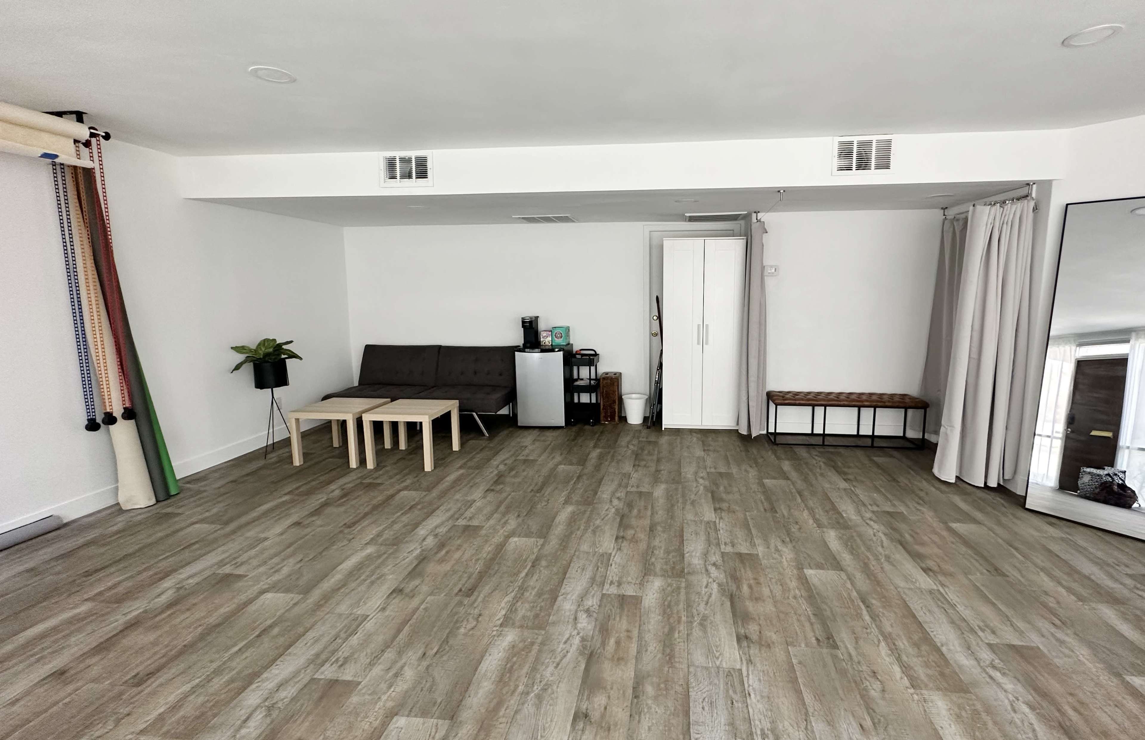 A spacious, light-filled room with wooden flooring, a sofa and coffee table, a small refrigerator, a cabinet, and a mirror along one wall.