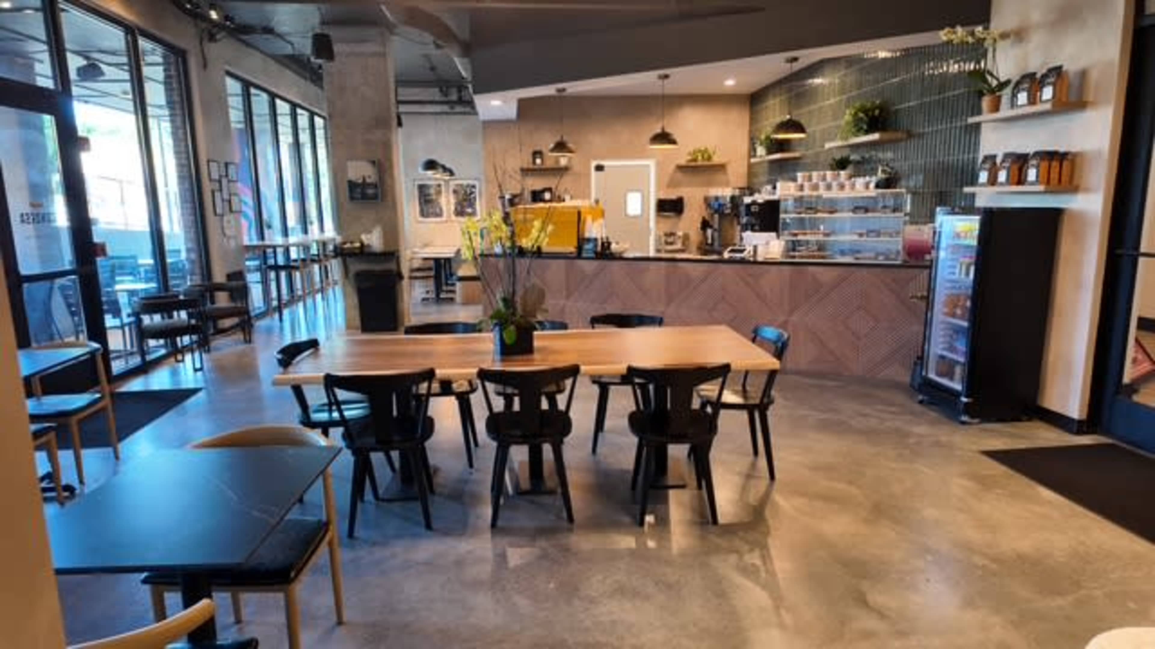 The image shows a modern café interior featuring a large wooden table, black chairs, a coffee counter, and large windows allowing natural light to enter.