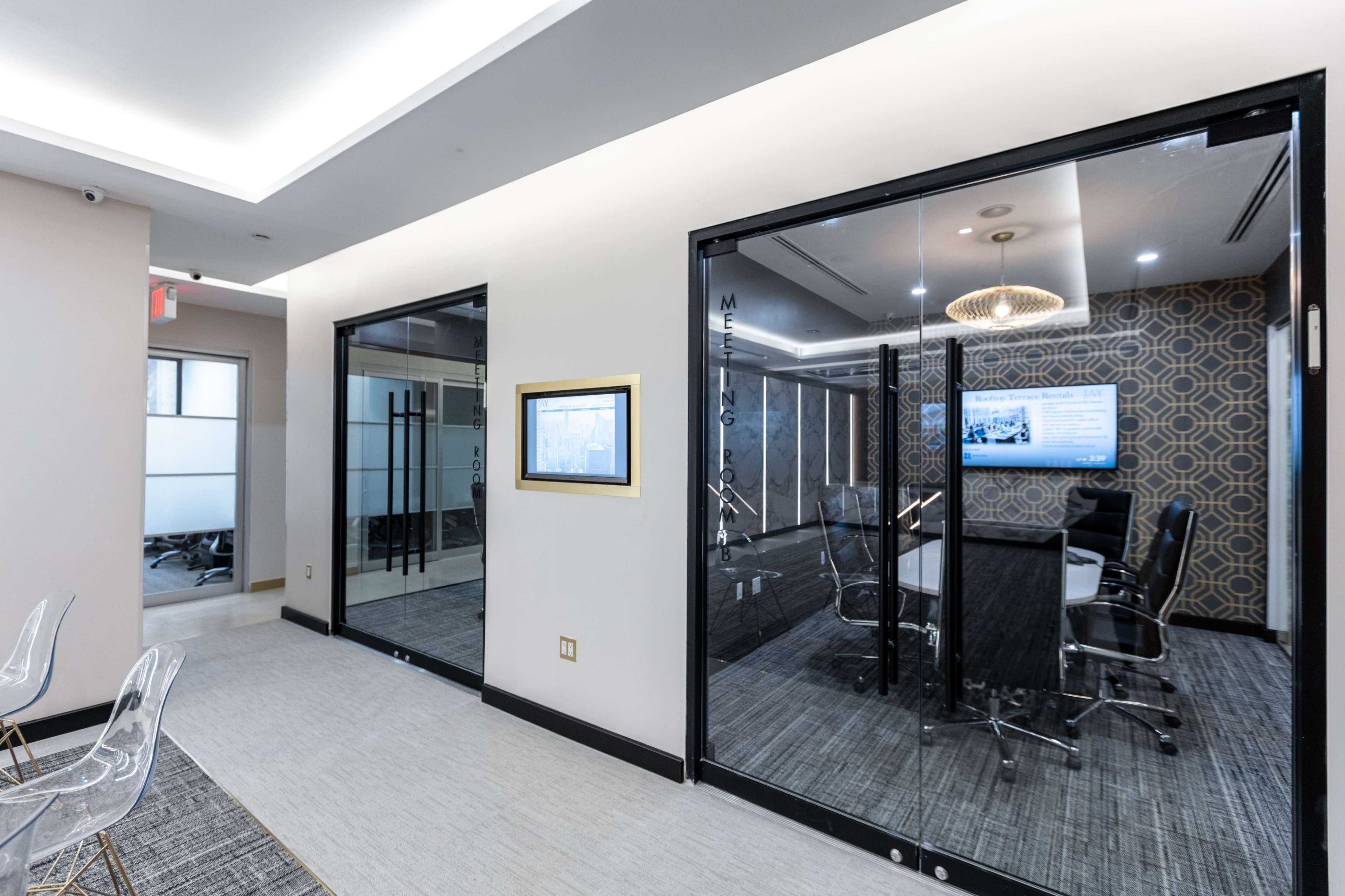 A modern office space featuring two glass-walled meeting rooms, each with a table and chairs, and a digital screen displaying information.