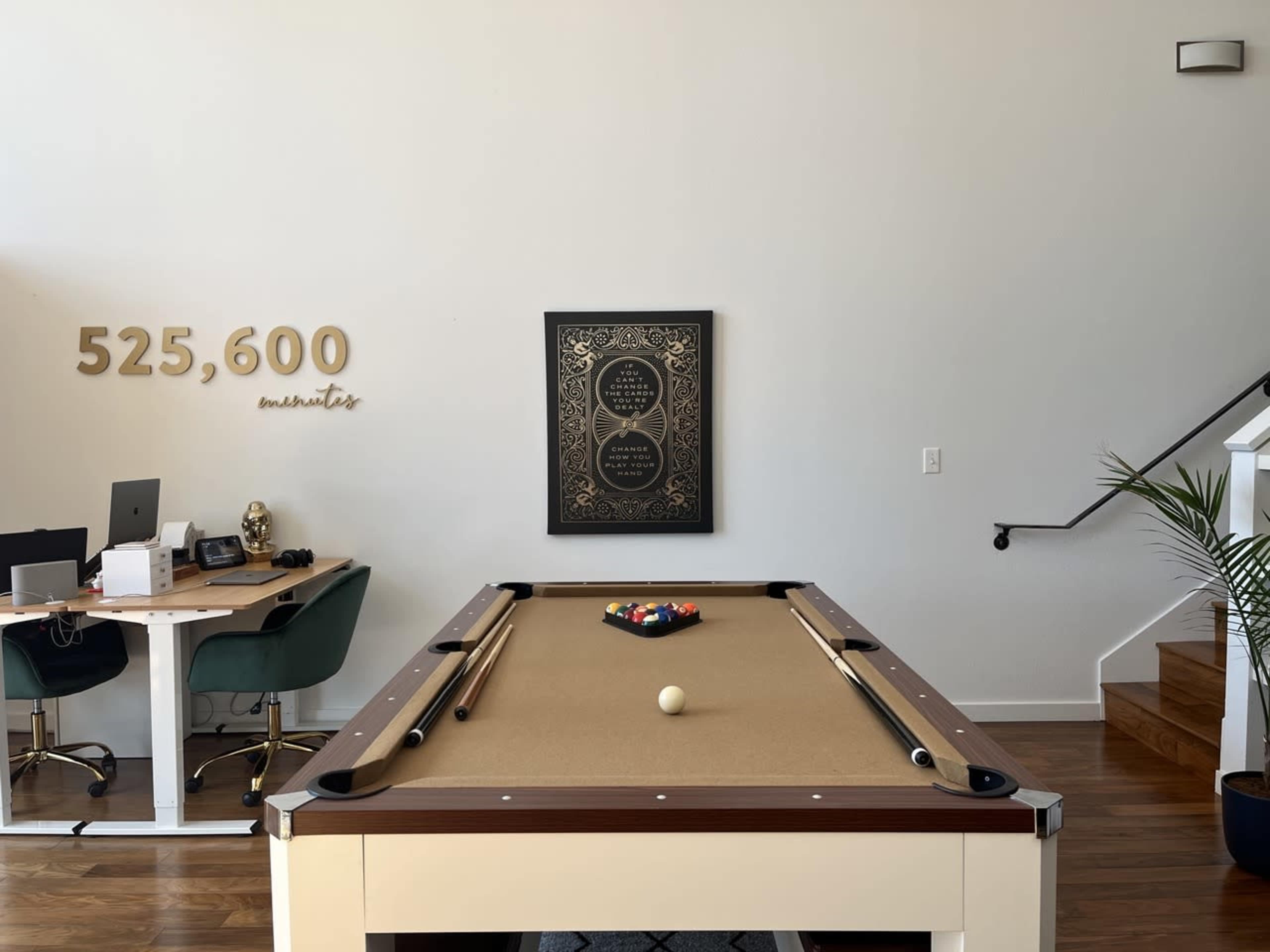 A pool table with a set of colored balls and cue sticks is positioned in a room with a desk and a framed art piece on the wall.