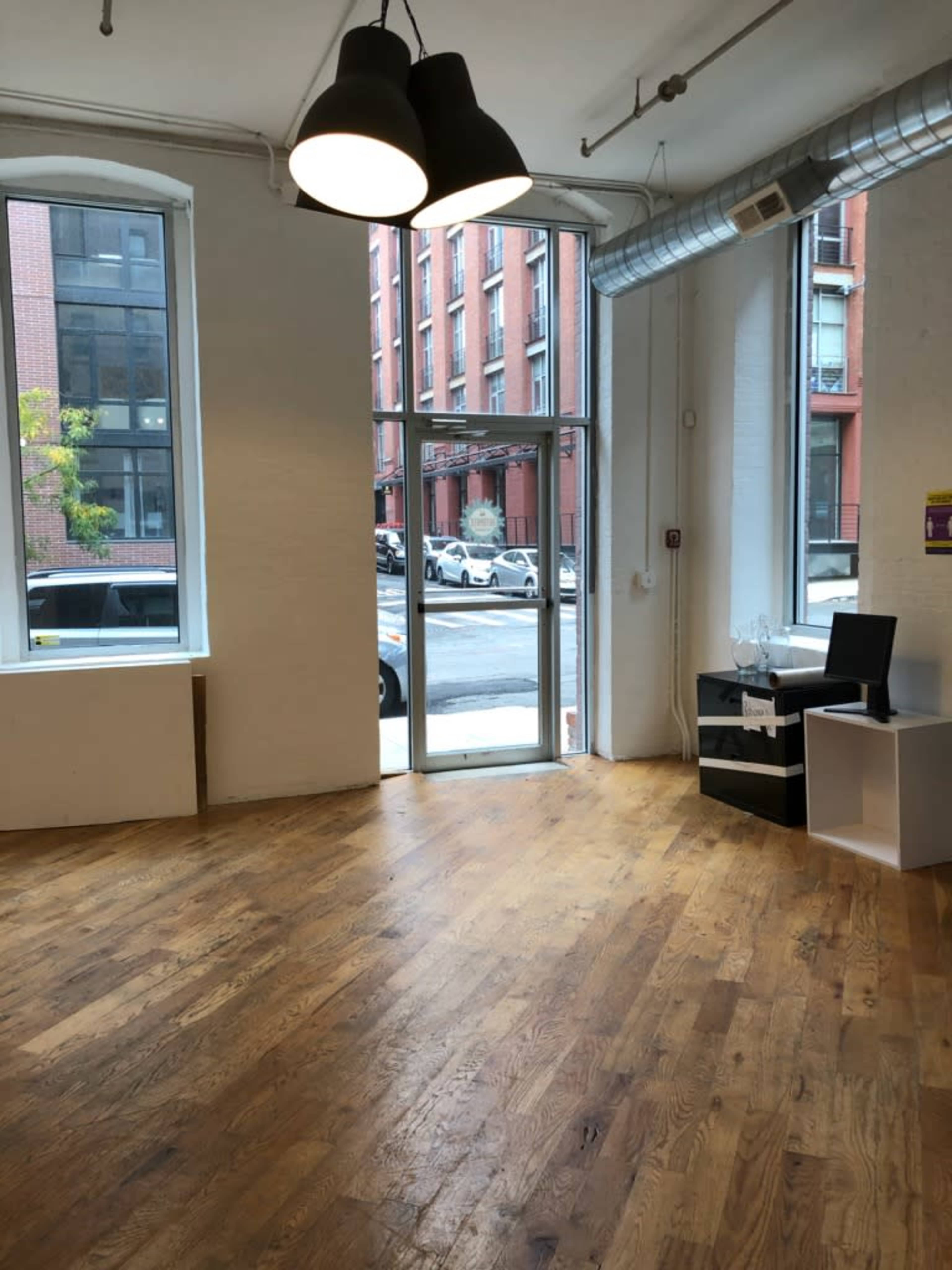 The image shows a spacious, empty room with large windows and wooden flooring, leading to an urban street view outside.