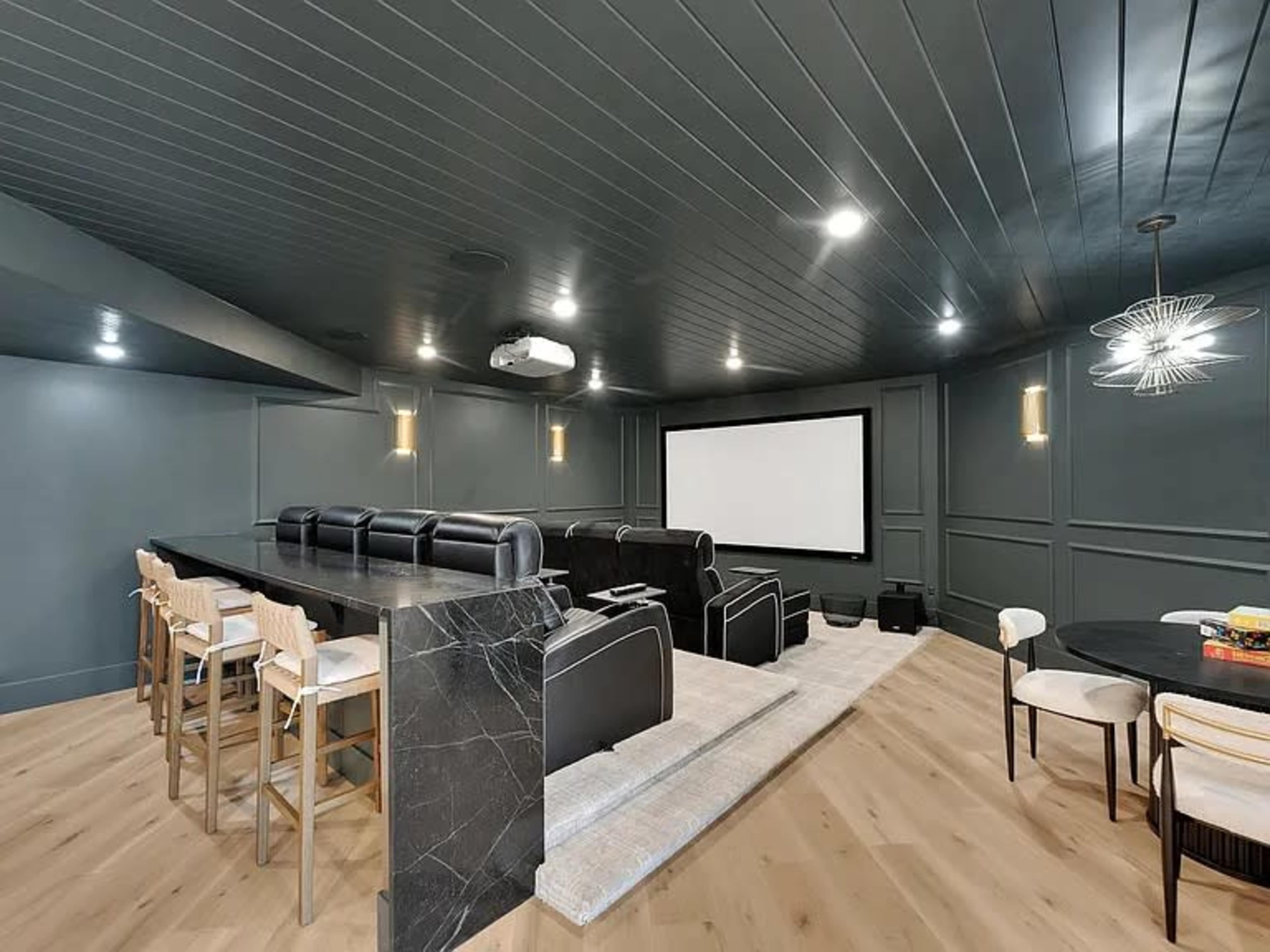 The image shows a home theater with a large screen, seating area with black chairs, a bar with stools, and a circular dining table, all set against dark walls and wood flooring.