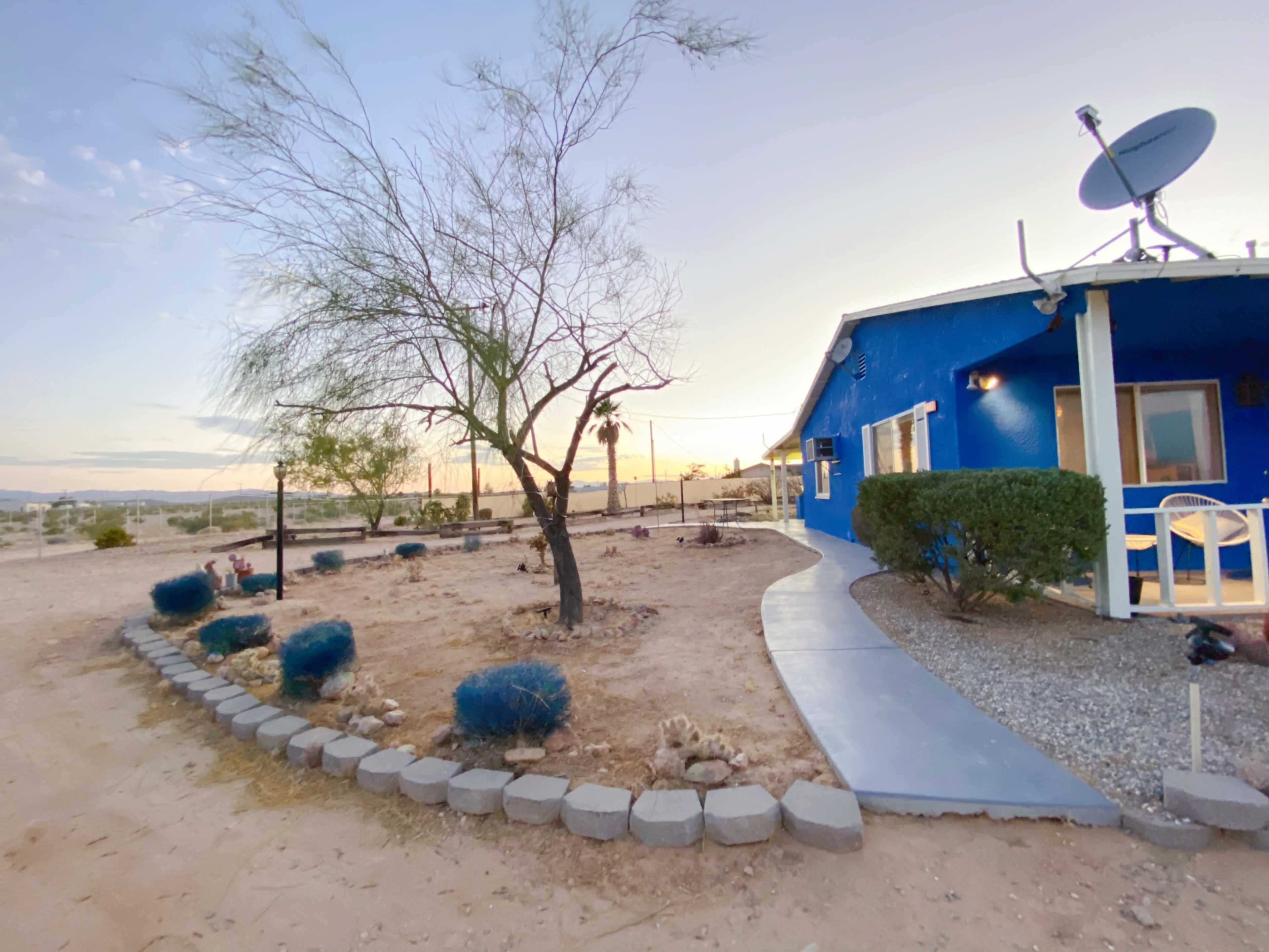 A blue house with a satellite dish sits at the edge of a desert landscape, surrounded by gravel and sparse vegetation under a twilight sky.