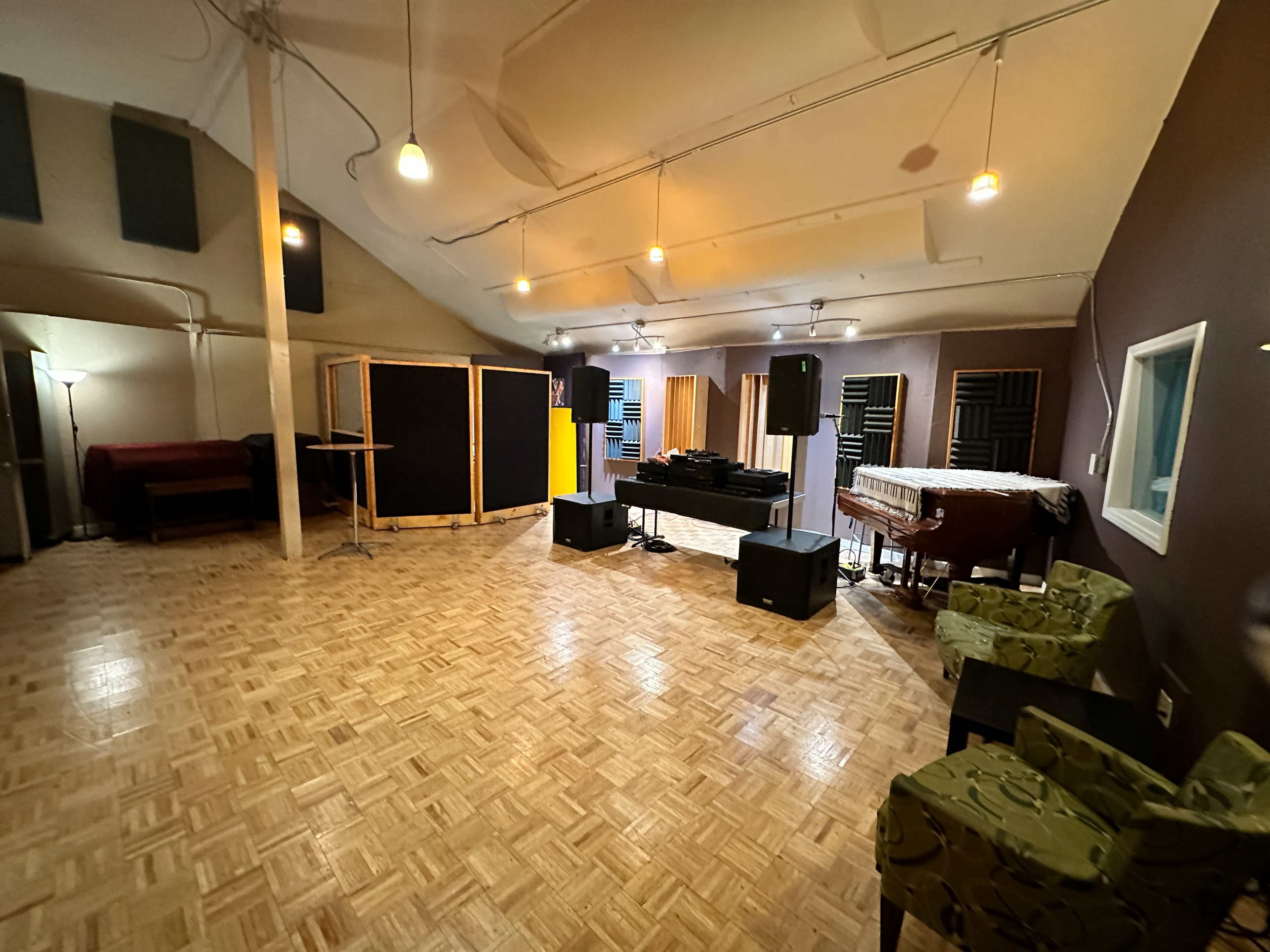 A spacious music studio with wooden flooring, soundproof booths, a table with equipment, and a couple of green chairs.