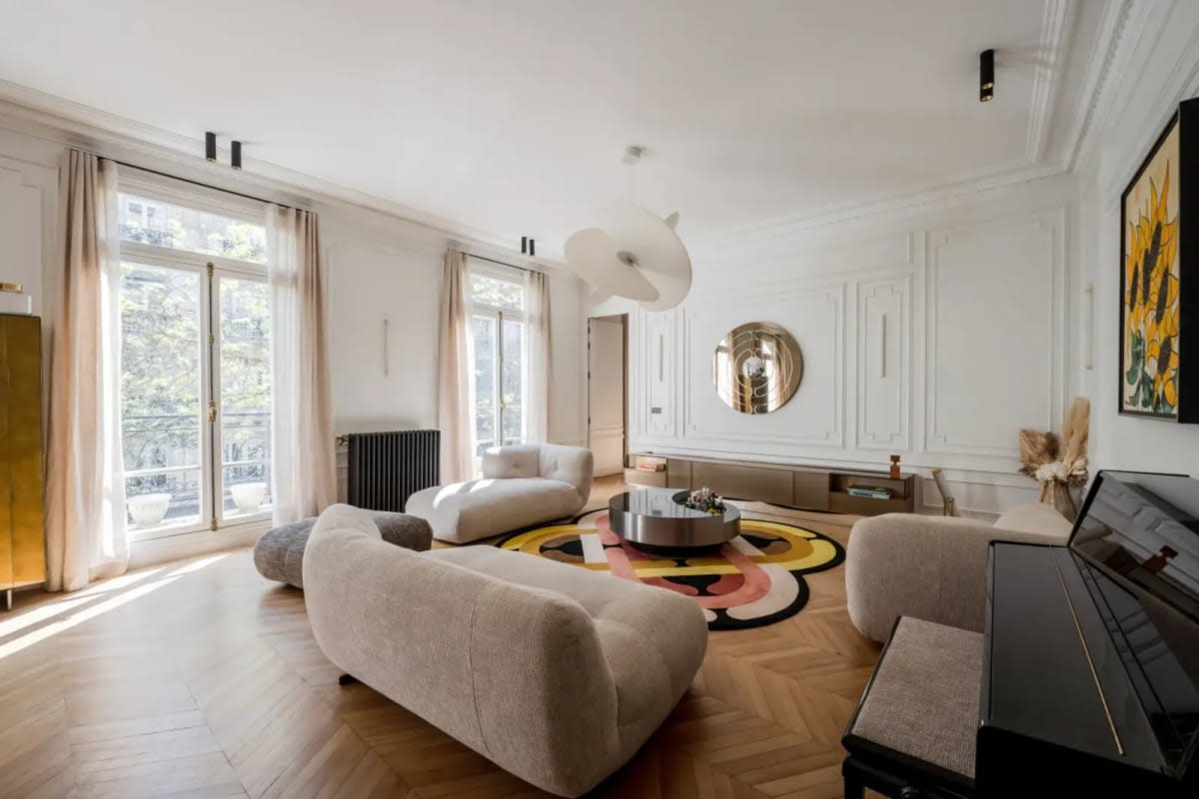 The room features a modern, spacious living area with a round coffee table, a large area rug, and large windows that allow natural light to enter.