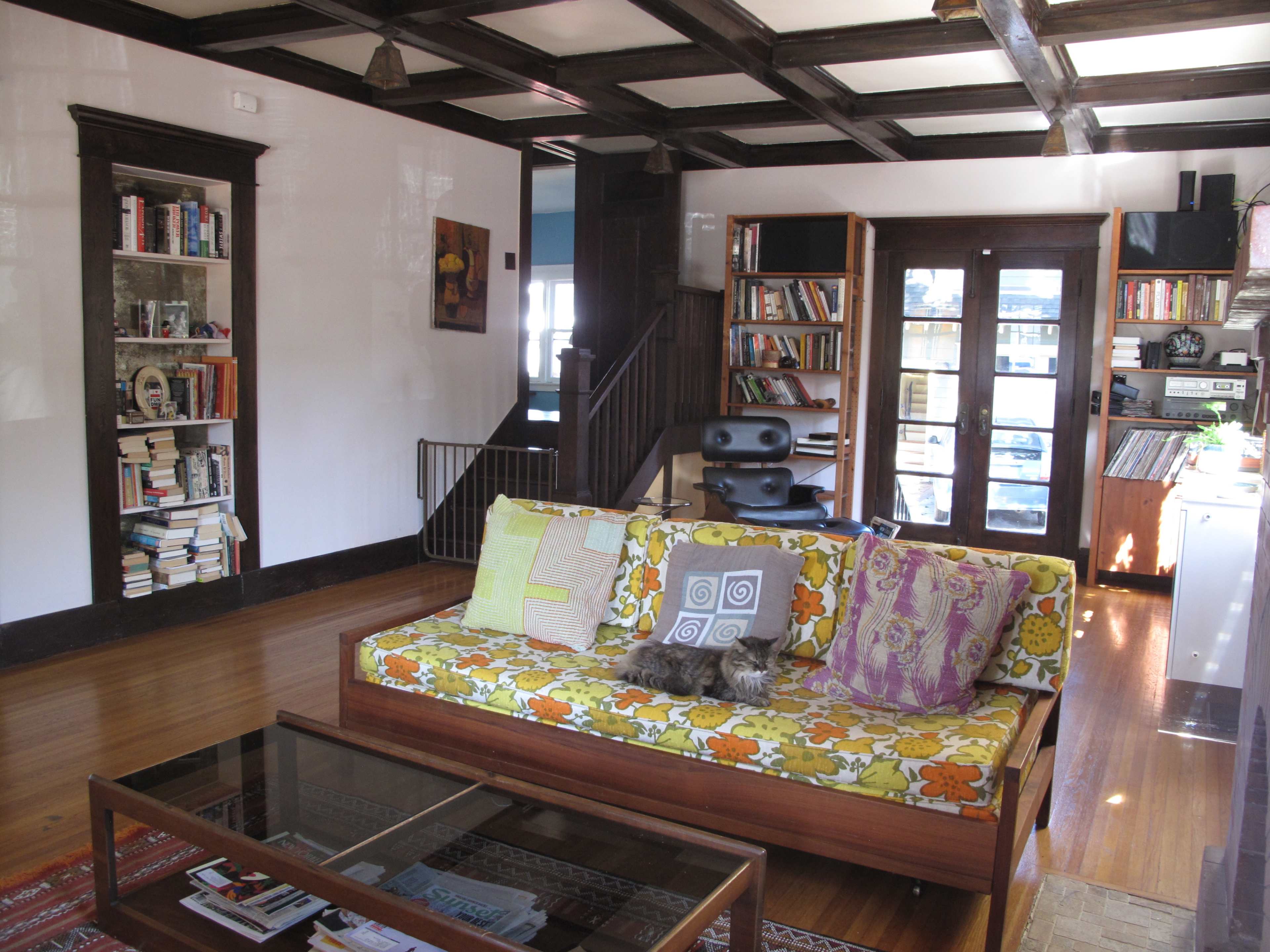 A cozy living room features a patterned couch, a coffee table, a bookshelf, and an open staircase leading to another level.