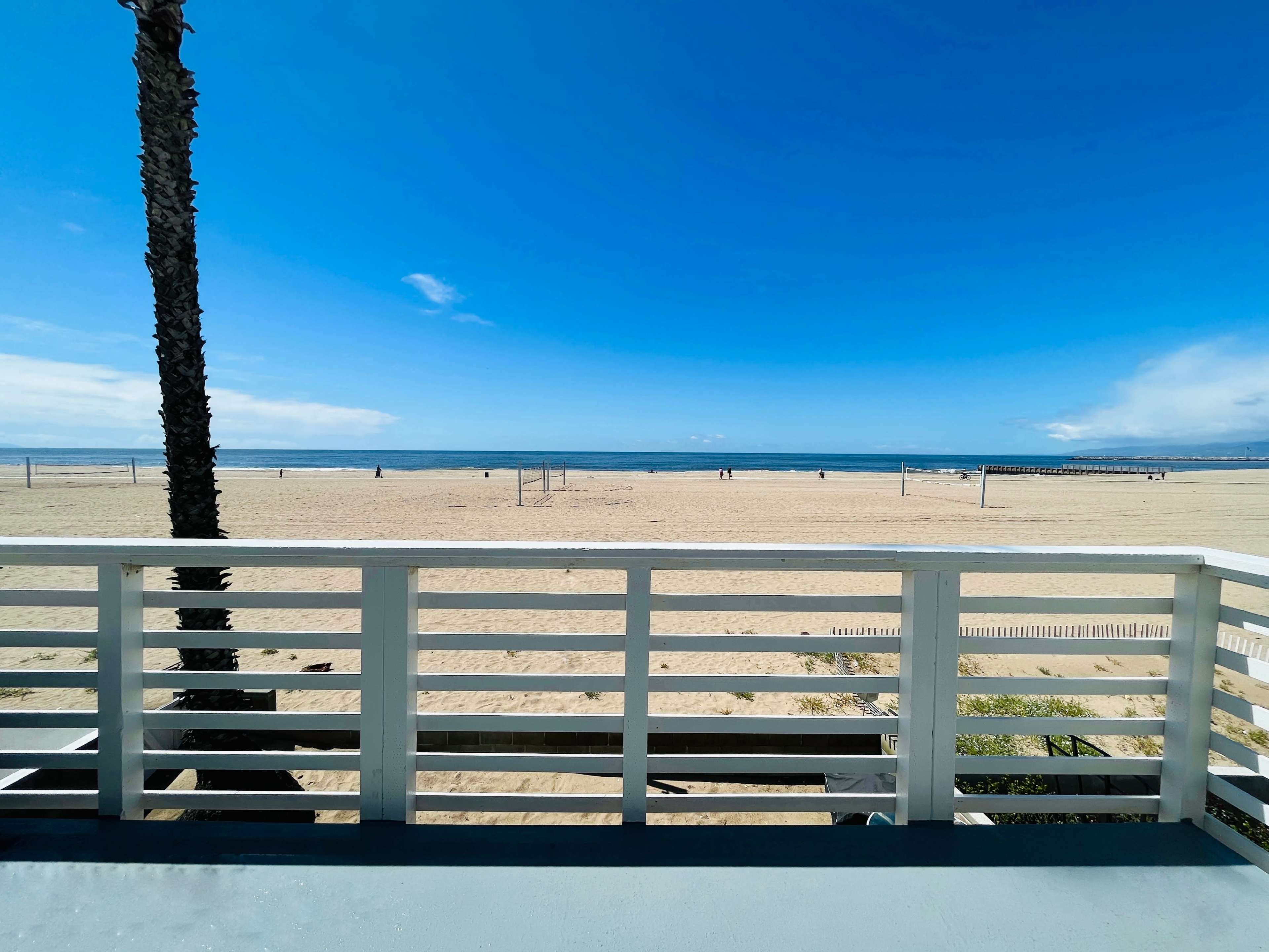 A view of a sandy beach with a blue sky and a few people in the distance, framed by a white railing and a palm tree.