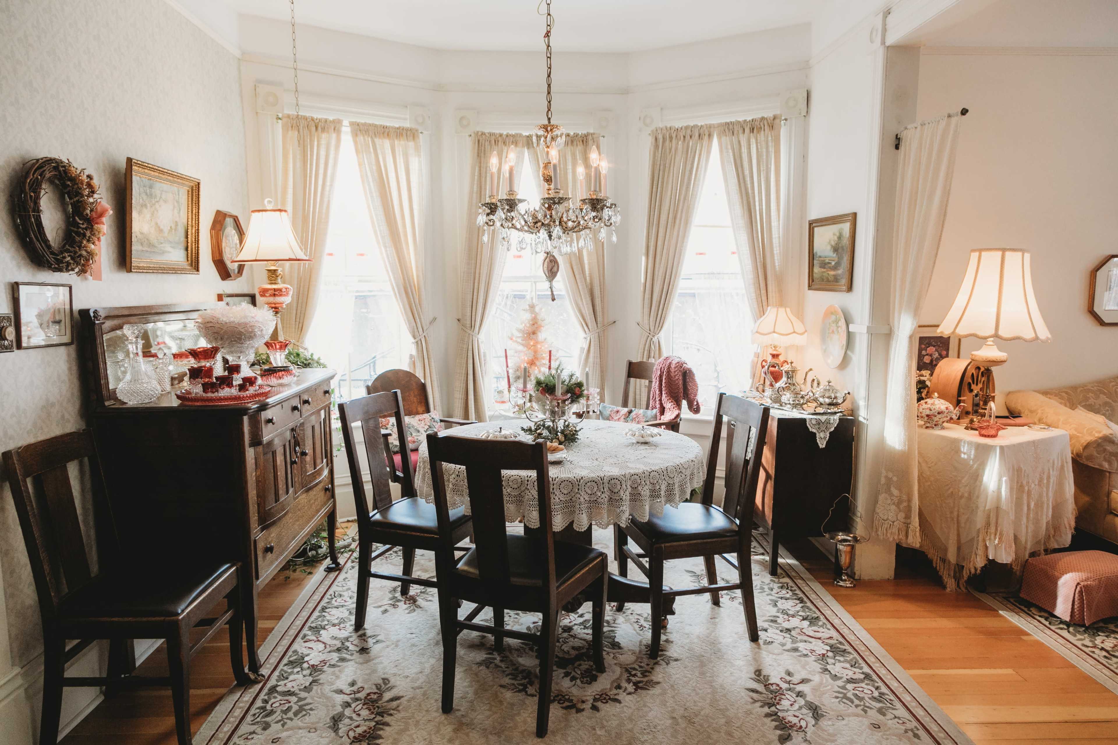 A dining room features a round table covered with a lace tablecloth, surrounded by wooden chairs, and is illuminated by a chandelier, with windows adorned by sheer curtains.