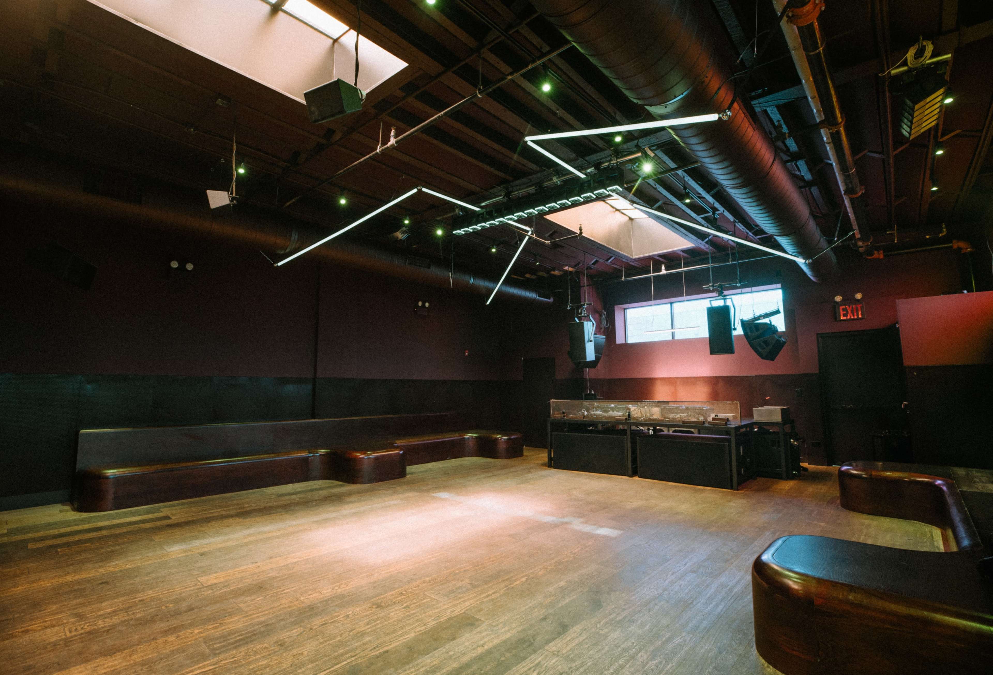 The image shows an empty interior of a dimly lit venue with wooden flooring and minimal seating along the walls, highlighted by angular lighting fixtures above.