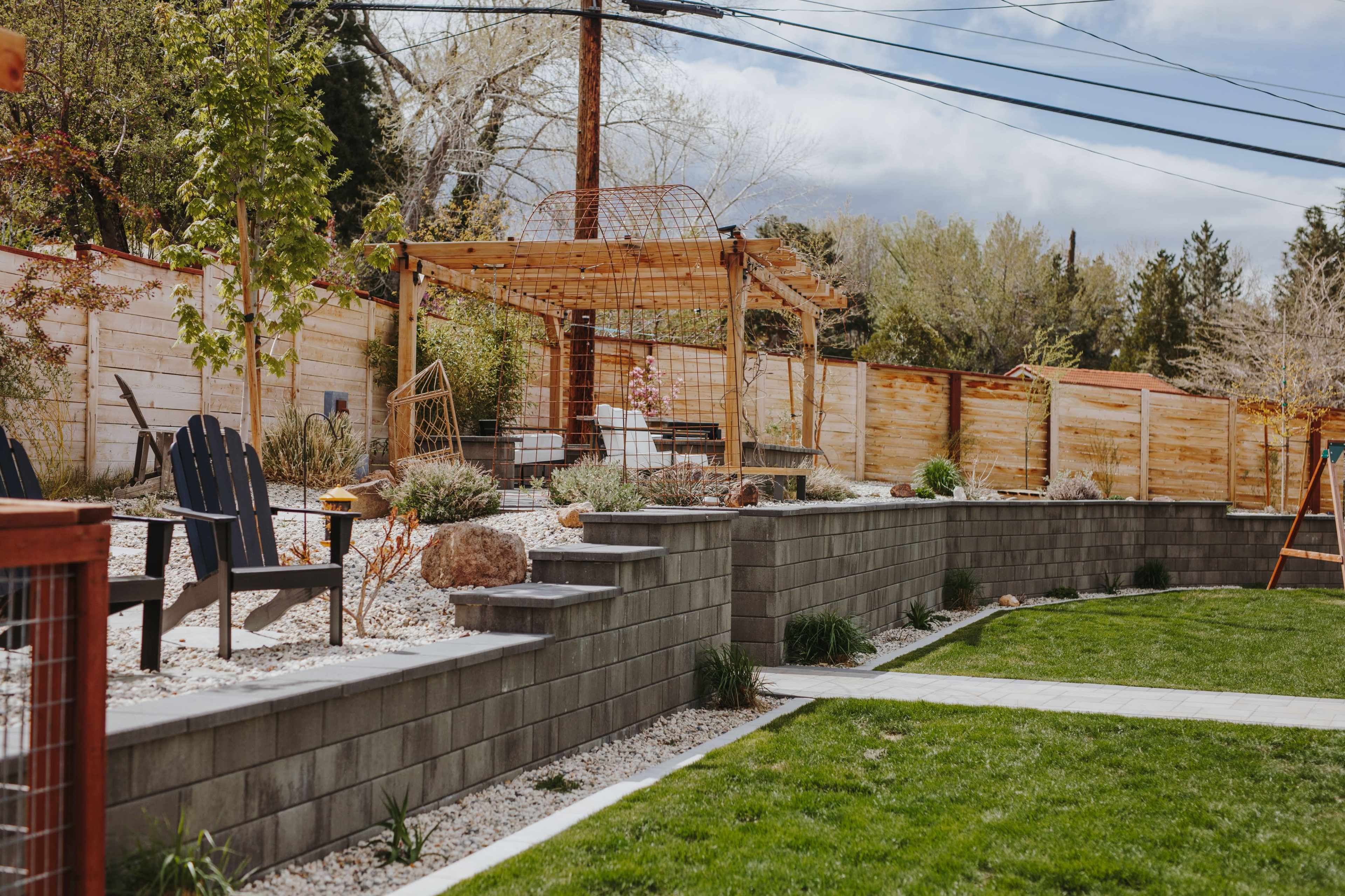A landscaped backyard with a stone wall, gravel beds, a pergola, and wooden seating areas surrounded by greenery.