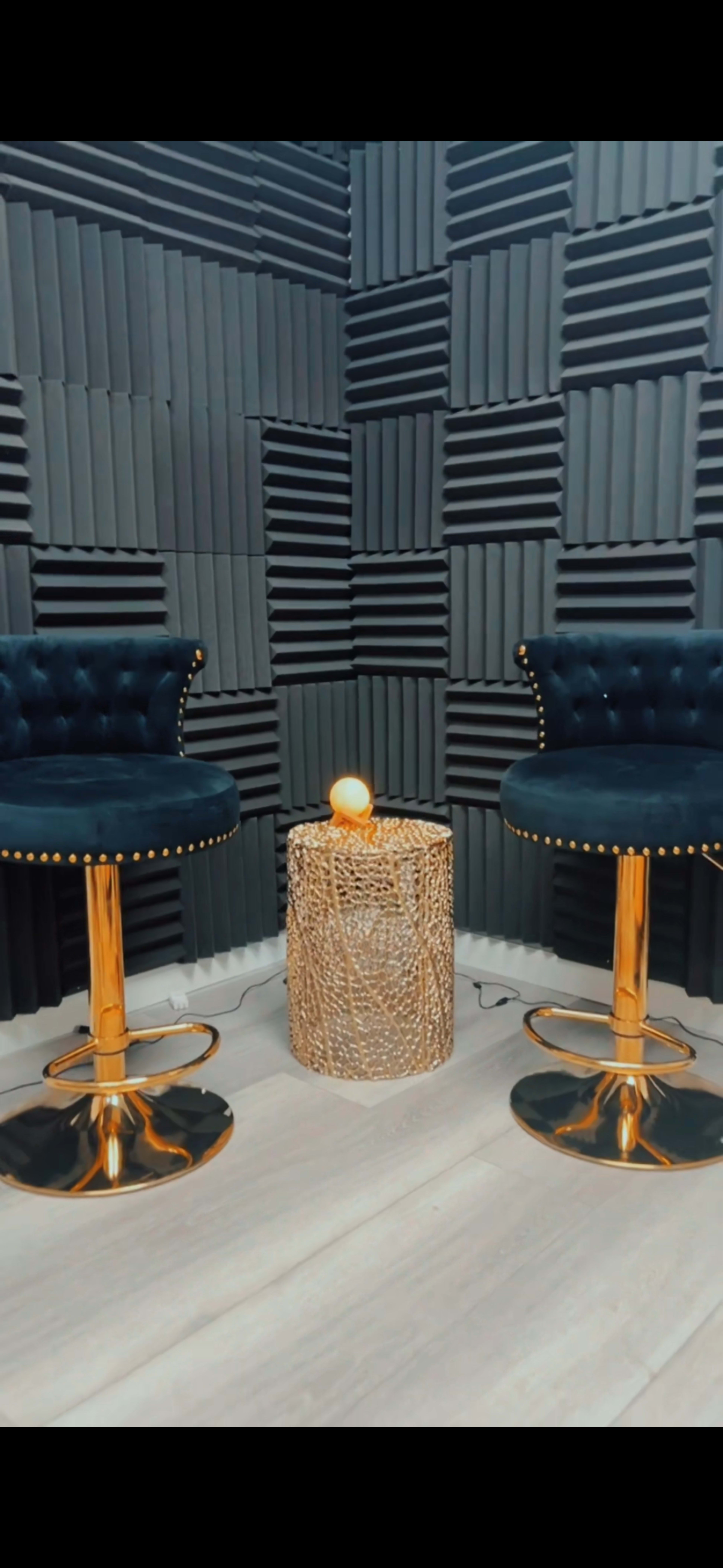 Two blue velvet bar stools with gold accents positioned on a light floor next to a woven, round table with a softly glowing orb on top, set against a wall of textured acoustic panels.