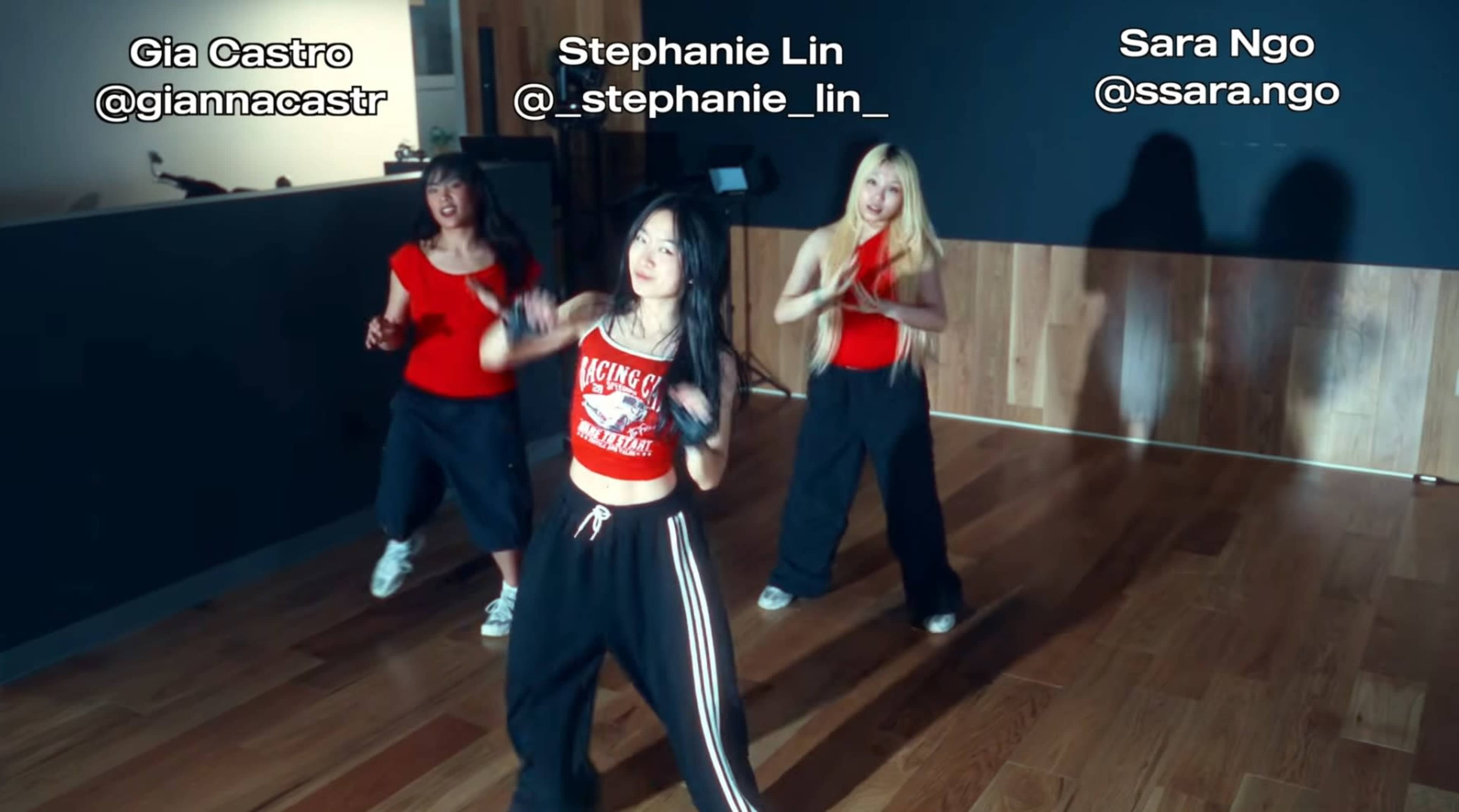 Three dancers perform in a studio with wooden flooring, wearing matching red and black outfits.