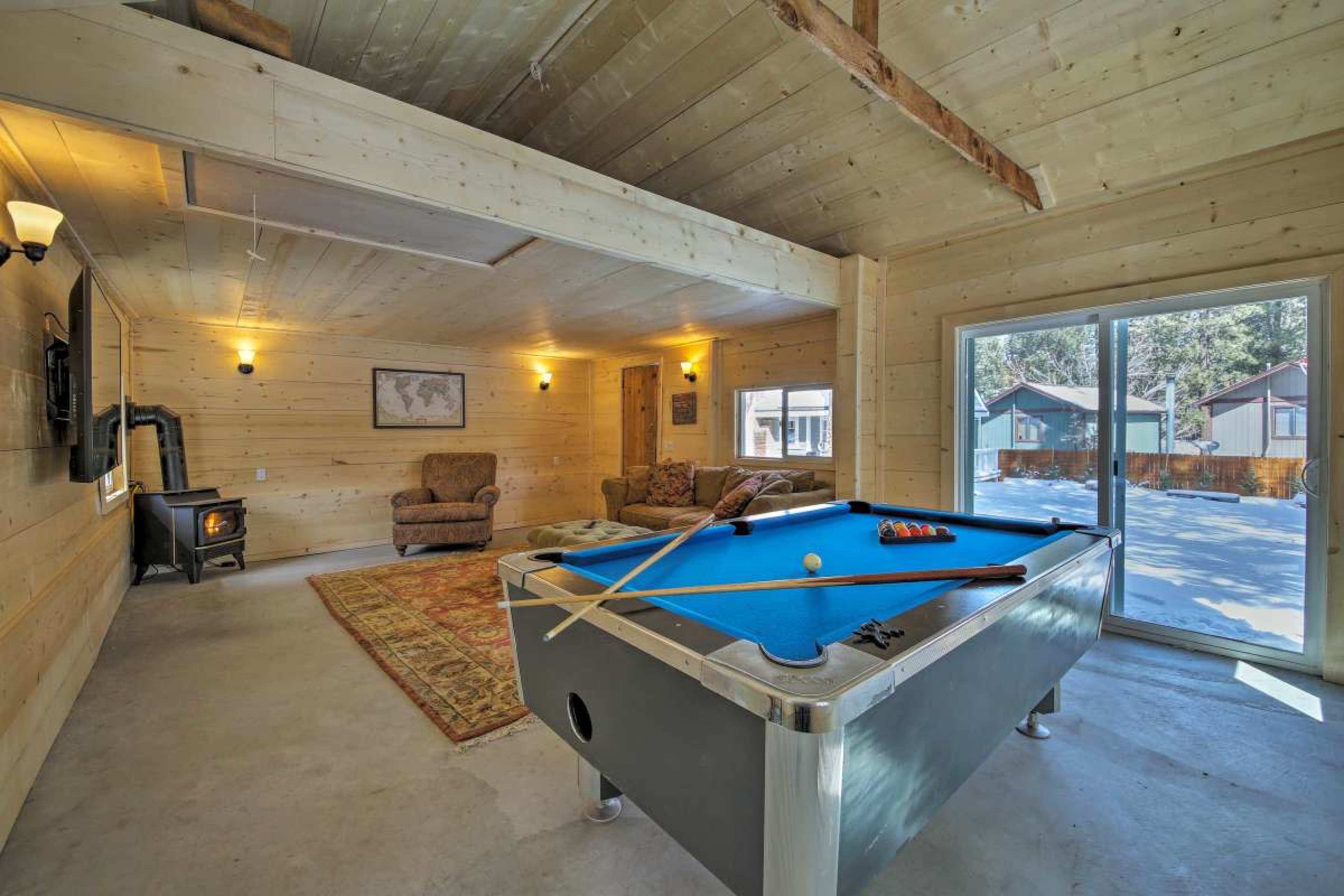 A light-colored wooden interior with a blue pool table, a sofa, and a wood stove, overlooking a snowy outdoor area through large glass doors.
