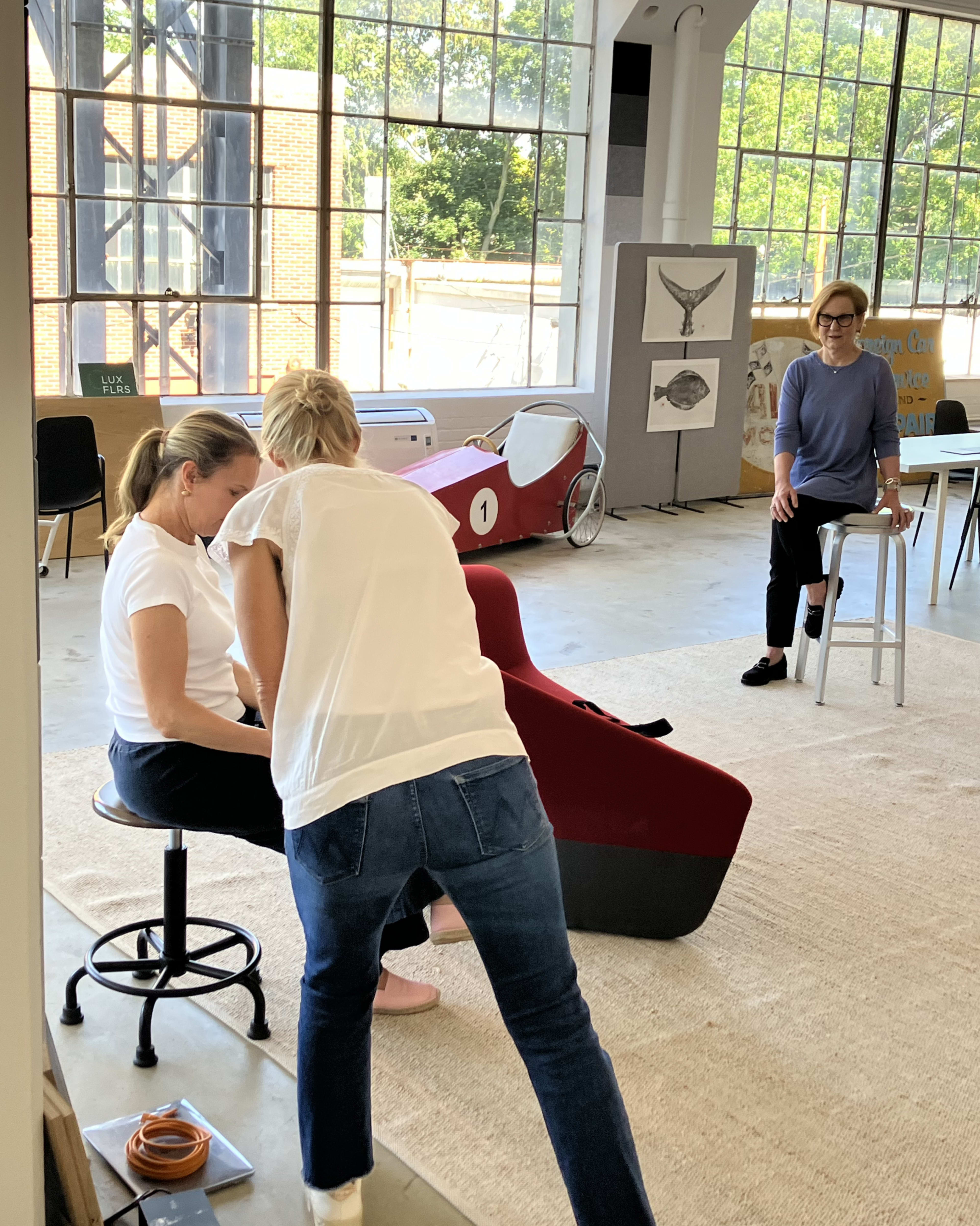 In a well-lit room with large windows, two women are engaged in a discussion while a third woman sits nearby on a stool.