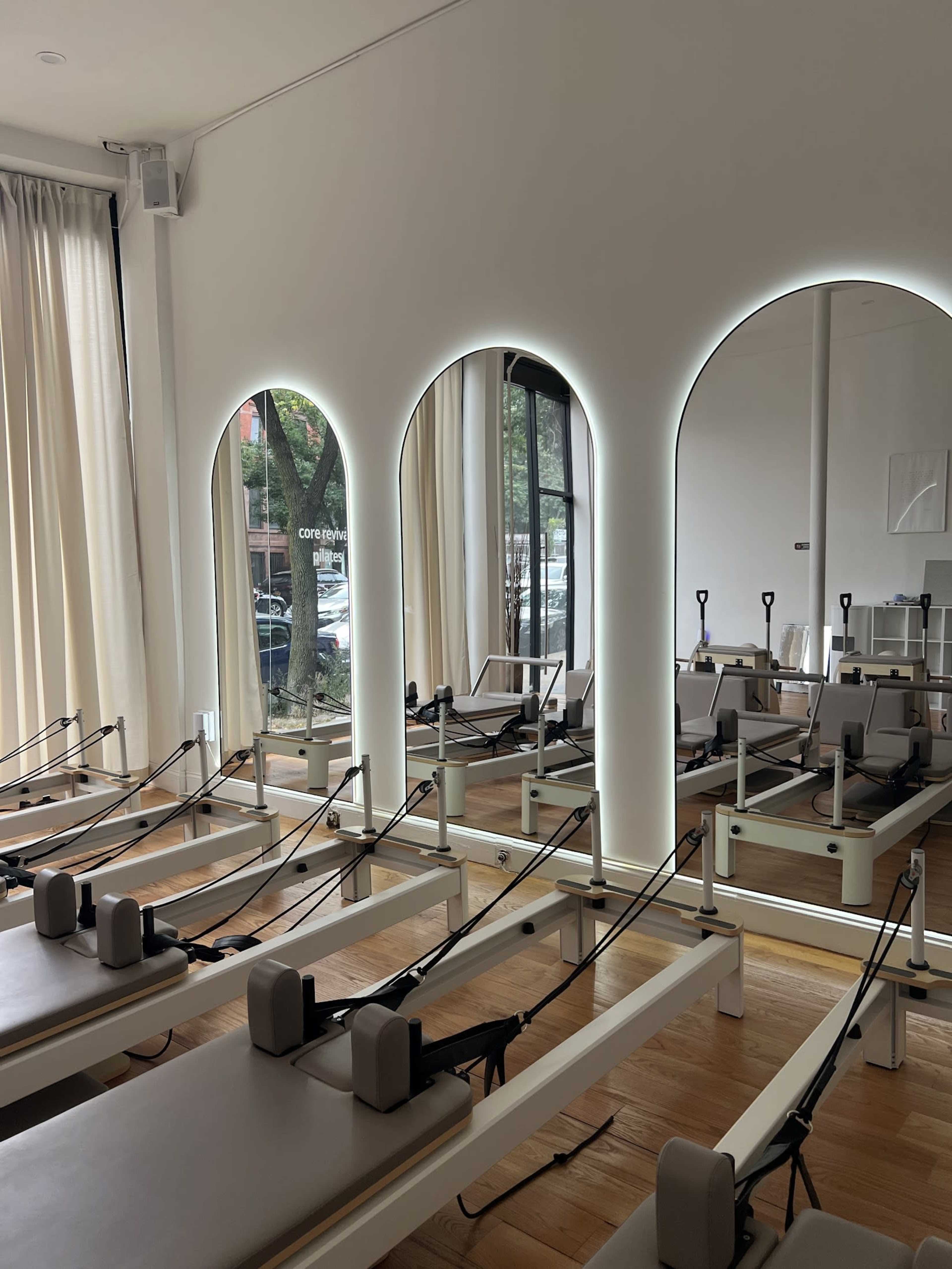 The image features a modern Pilates studio with reformers arranged neatly in rows, illuminated by arched mirrors with backlighting.