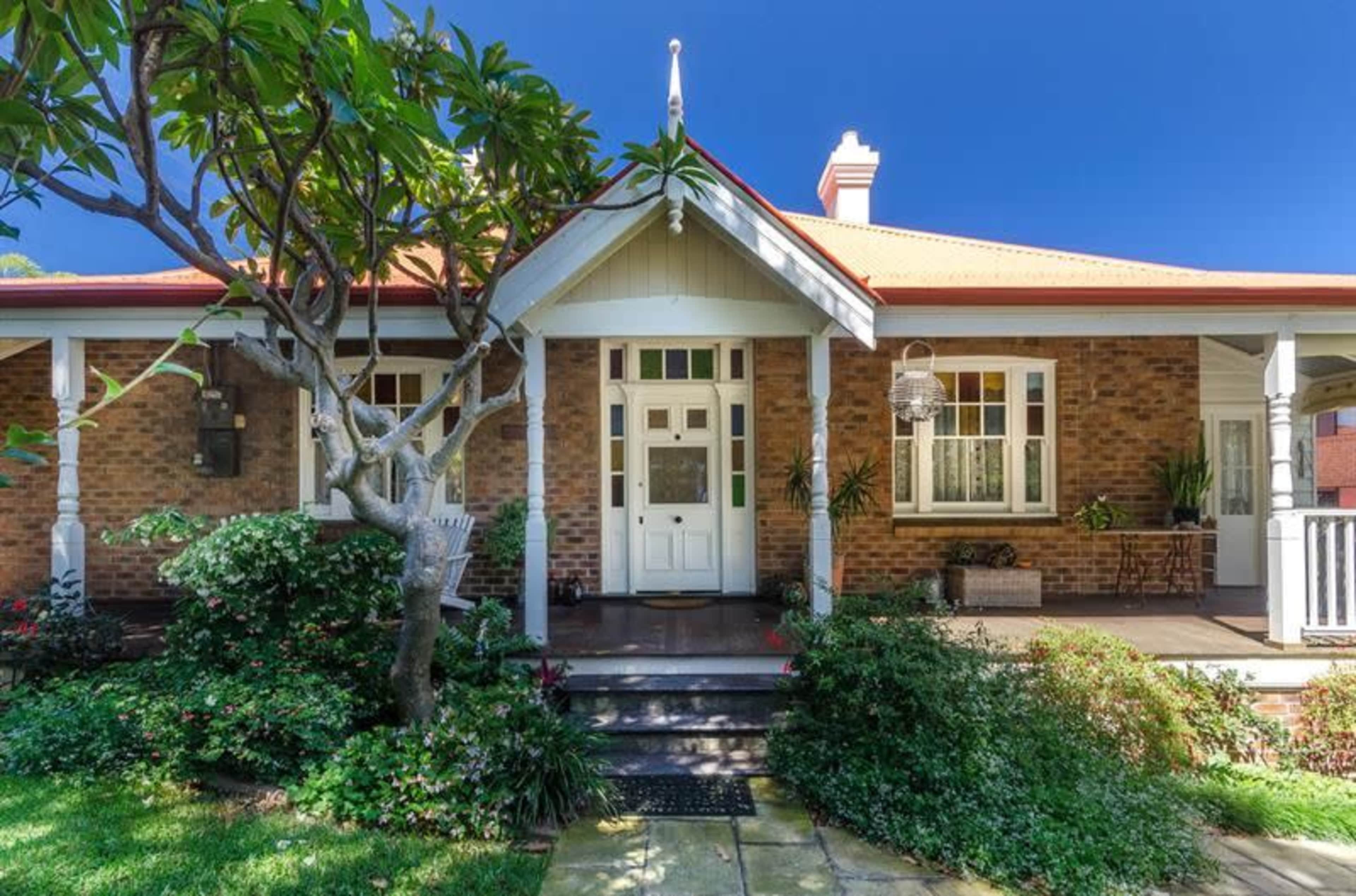 The image shows a single-story brick house with a front porch, surrounded by greenery and a well-maintained garden.