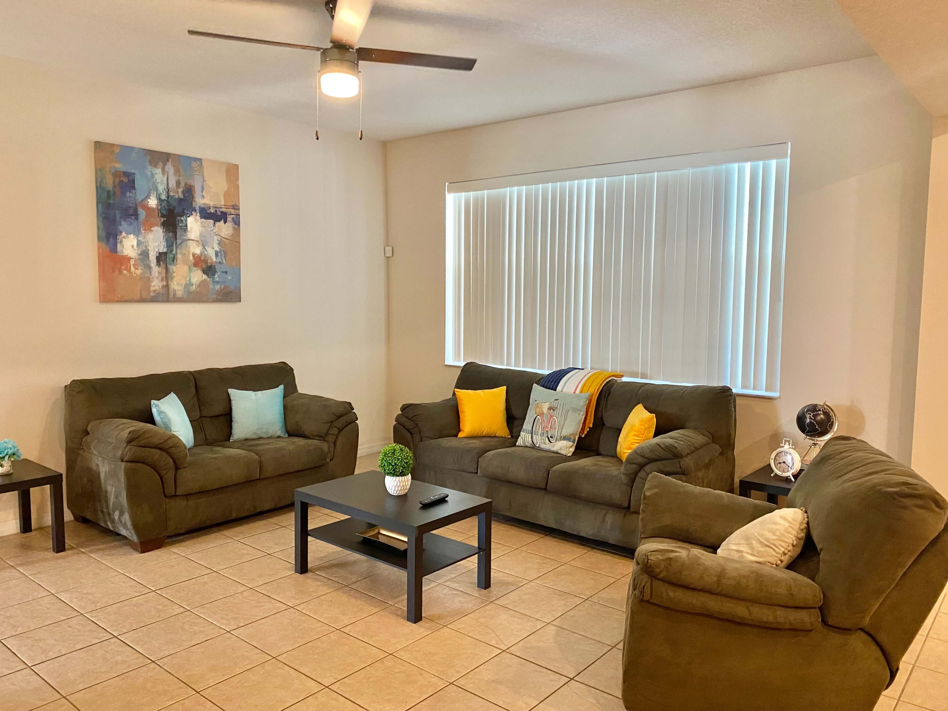 A living room features three green couches, a coffee table, a ceiling fan, and a large window with blinds.