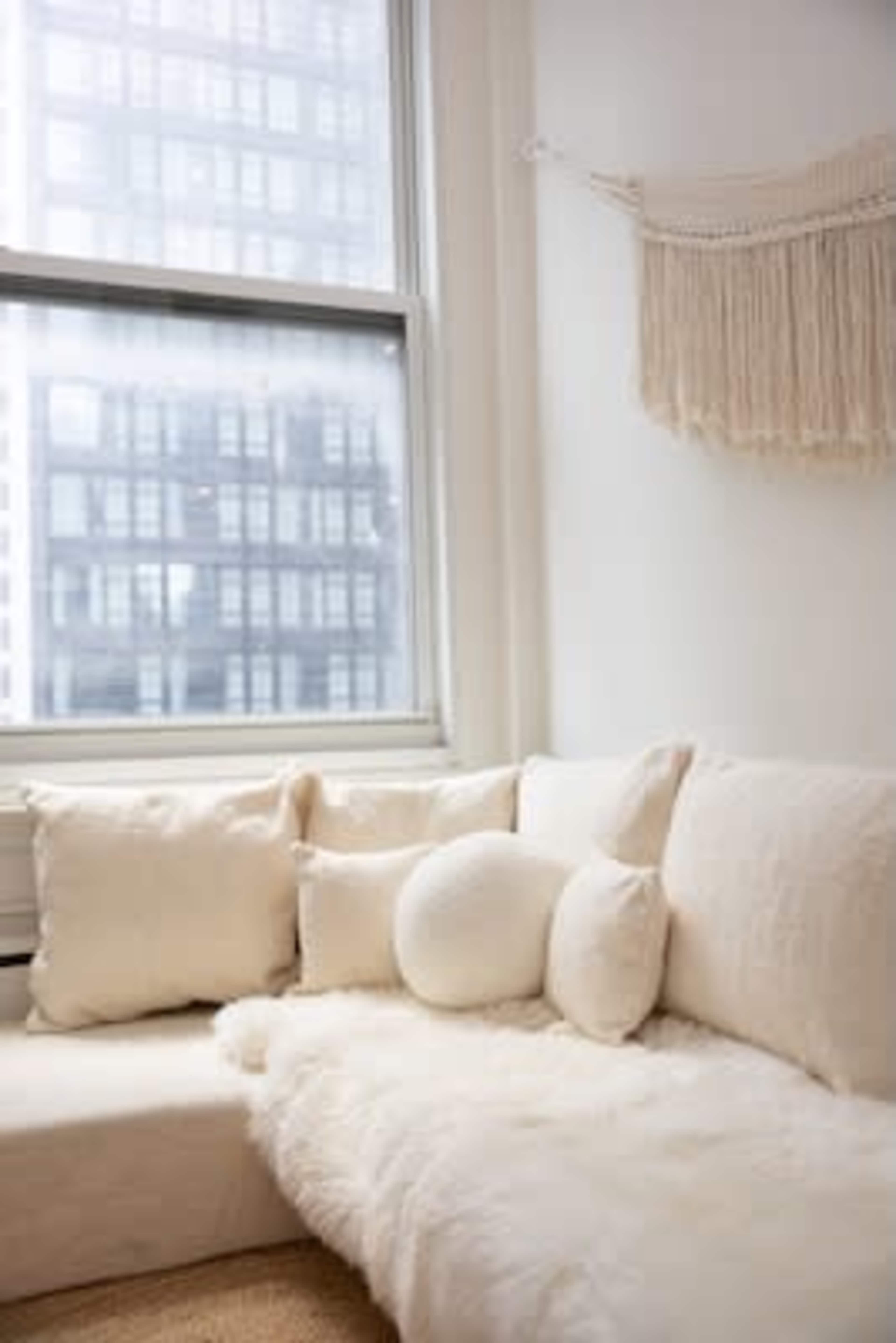 The image shows a cozy seating area with a white couch adorned with several cream-colored pillows and a fluffy throw, against a backdrop of a window revealing a cityscape.