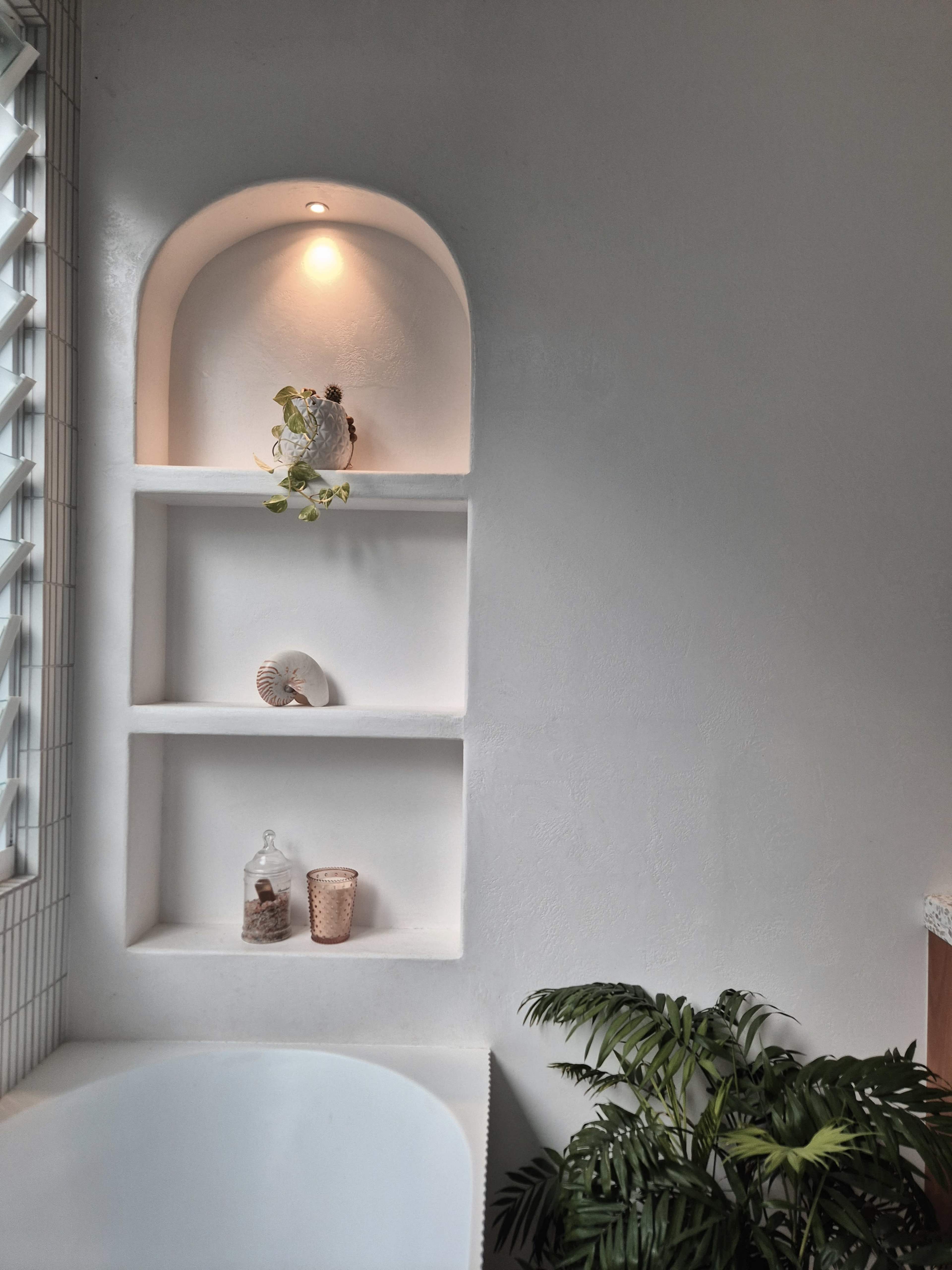 The image shows a minimalist bathroom with white walls, built-in shelves displaying decor items, and a small green plant in the corner.