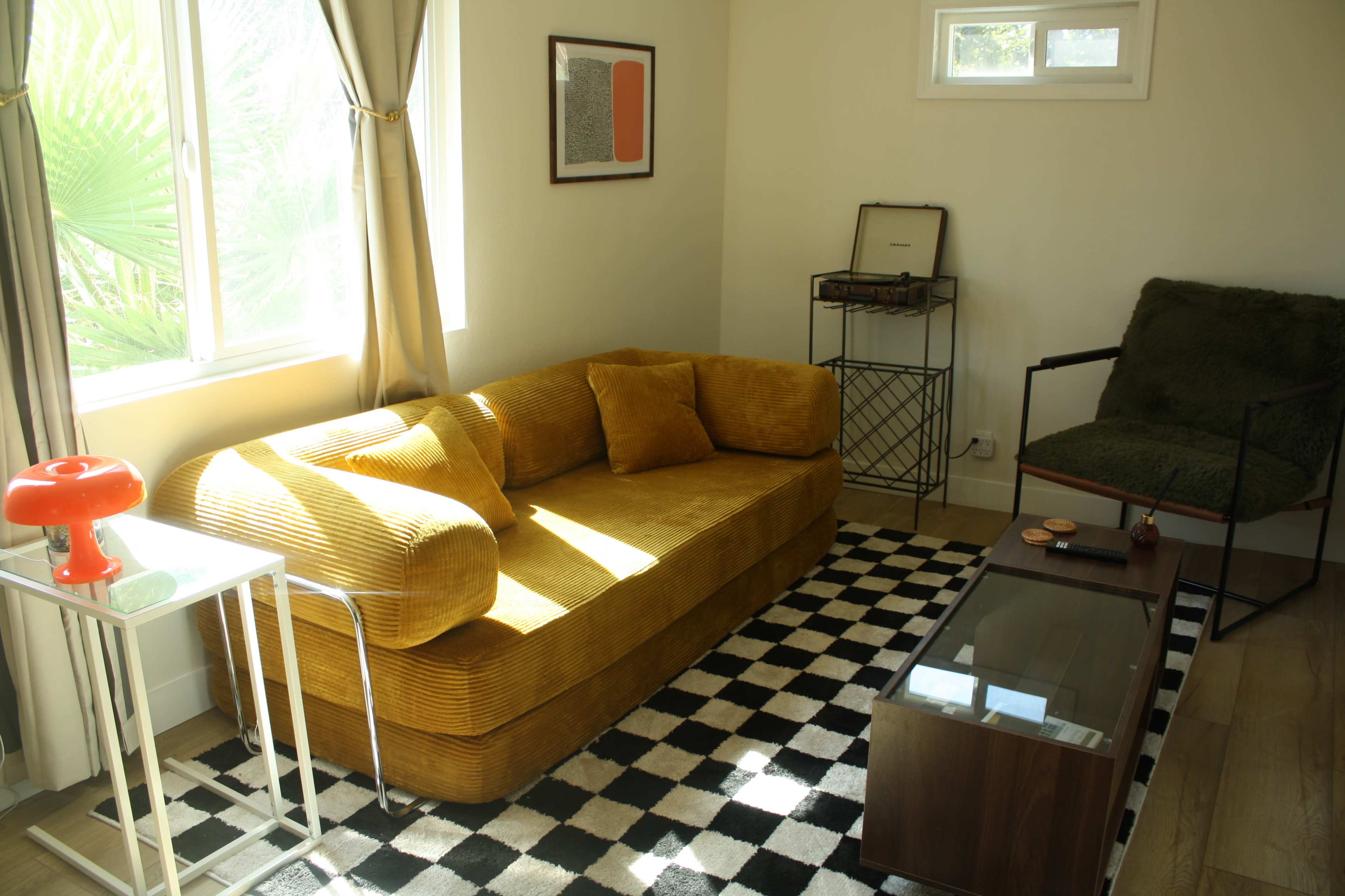 A yellow corduroy sofa and a black-and-white checkered rug are presented in a bright living room with a record player, a side table, and a green armchair.