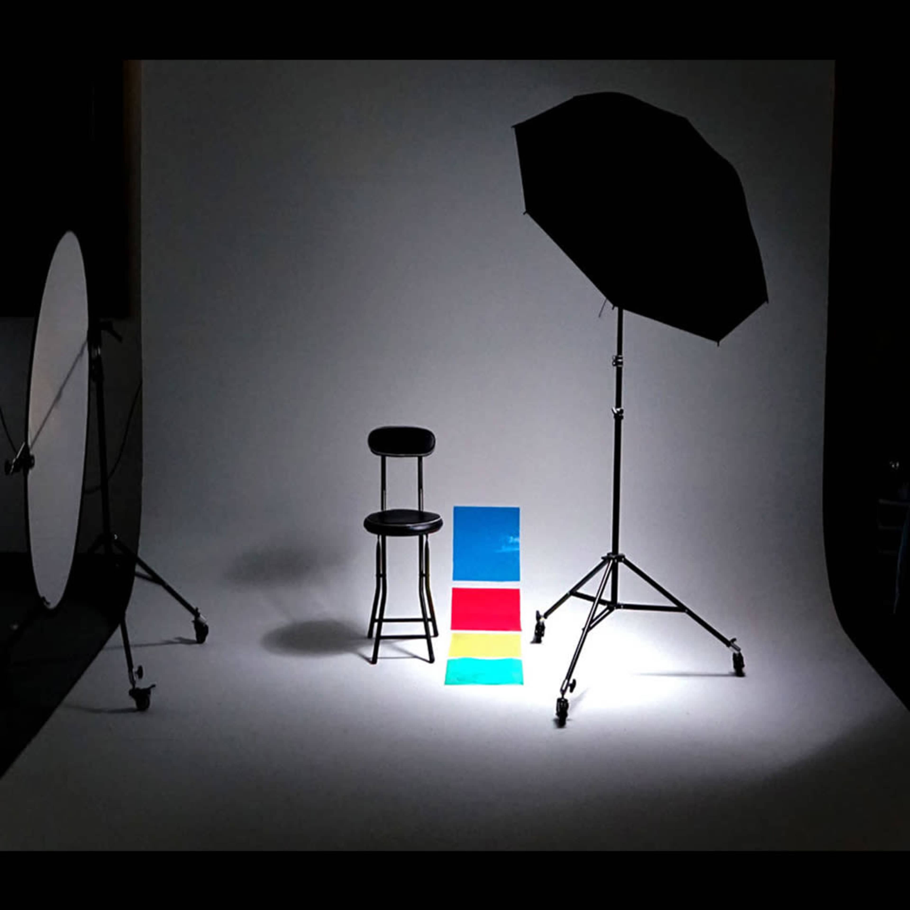 The image shows a photography setup with a black chair positioned in front of a white backdrop, along with colored panels and a large umbrella light.