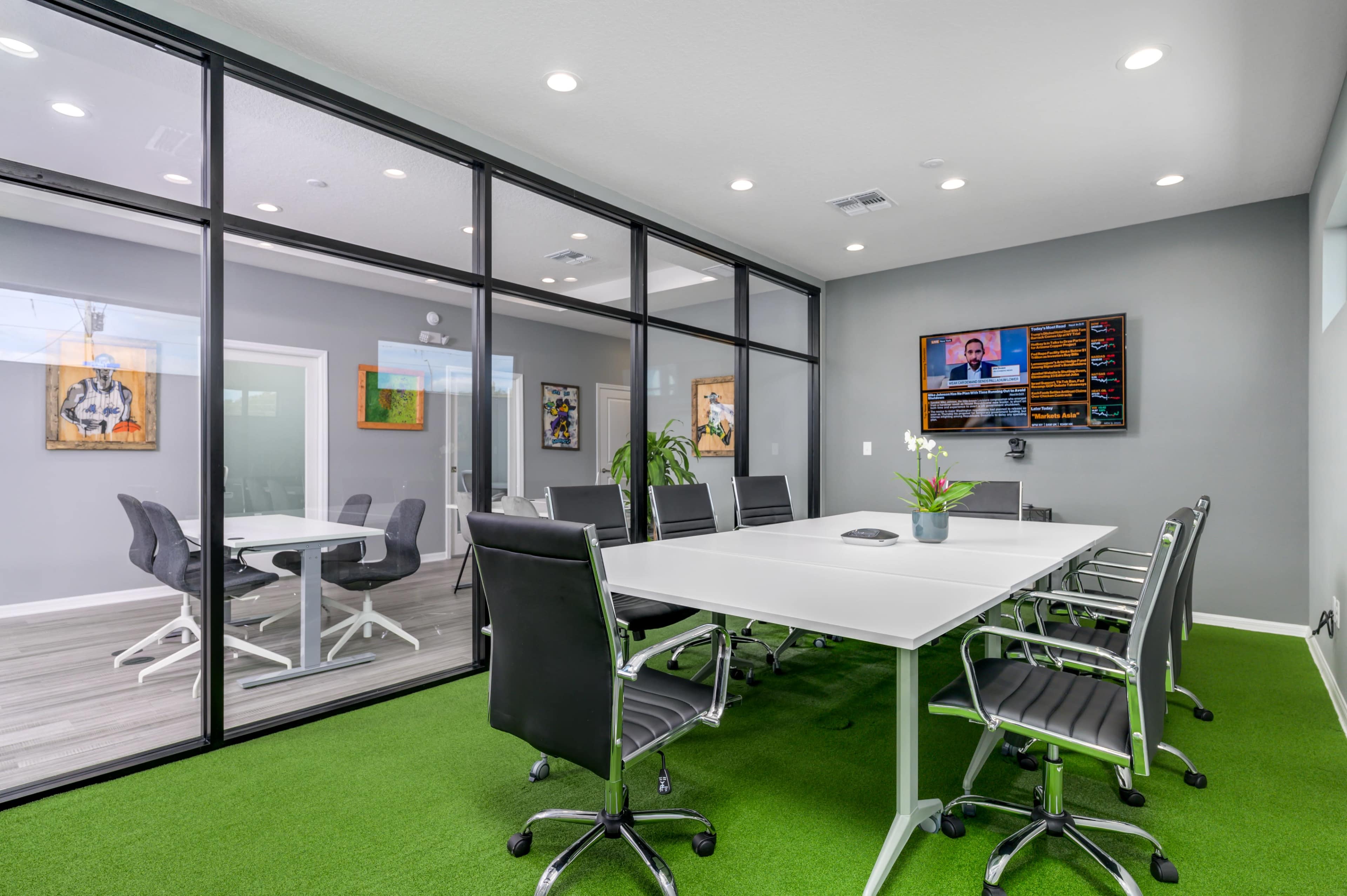 A modern conference room features a large white table surrounded by black chairs, with glass walls and a television displaying information on one side.