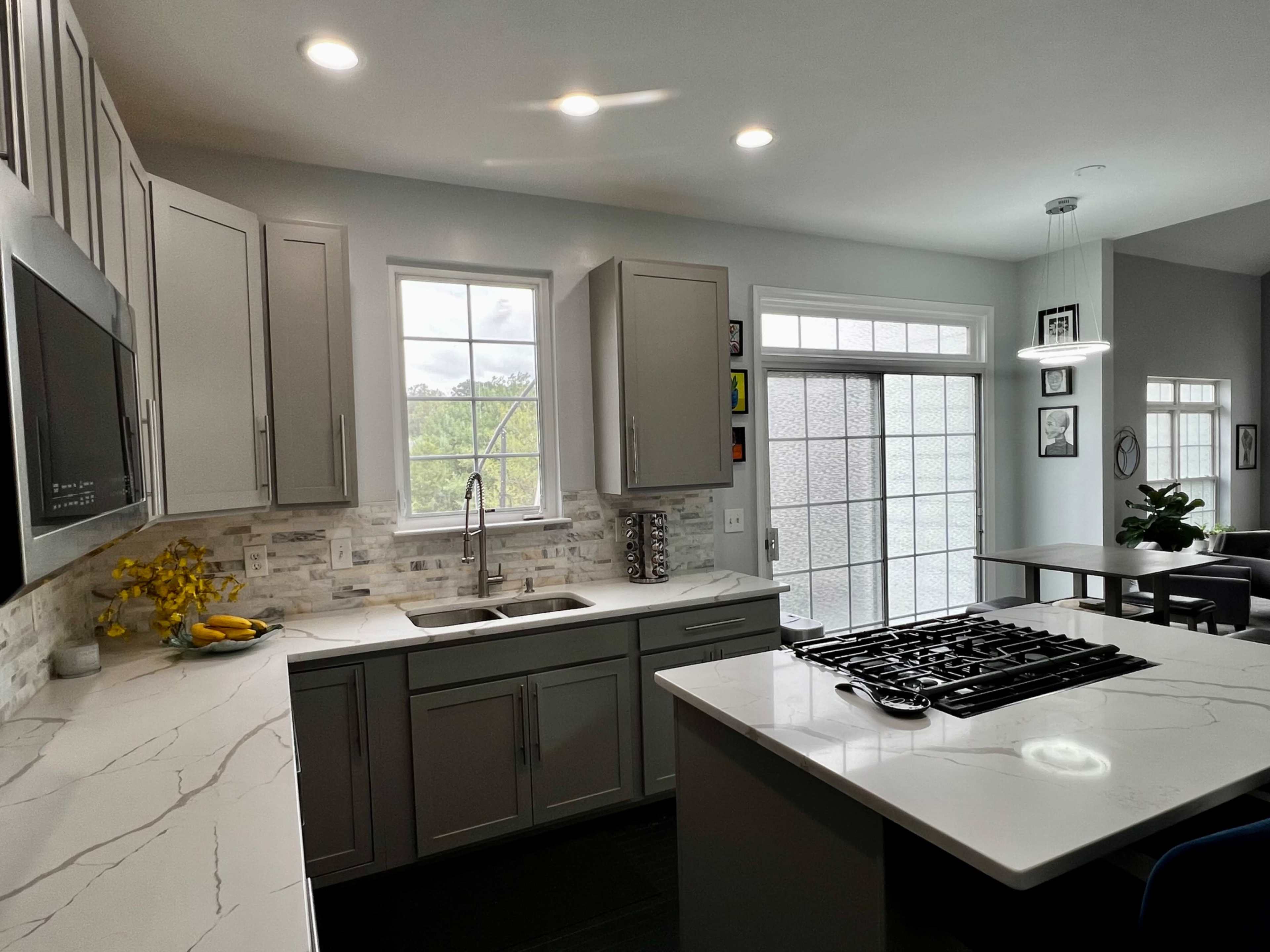 A modern kitchen features gray cabinets, a white countertop with a stovetop, and large windows that provide natural light.