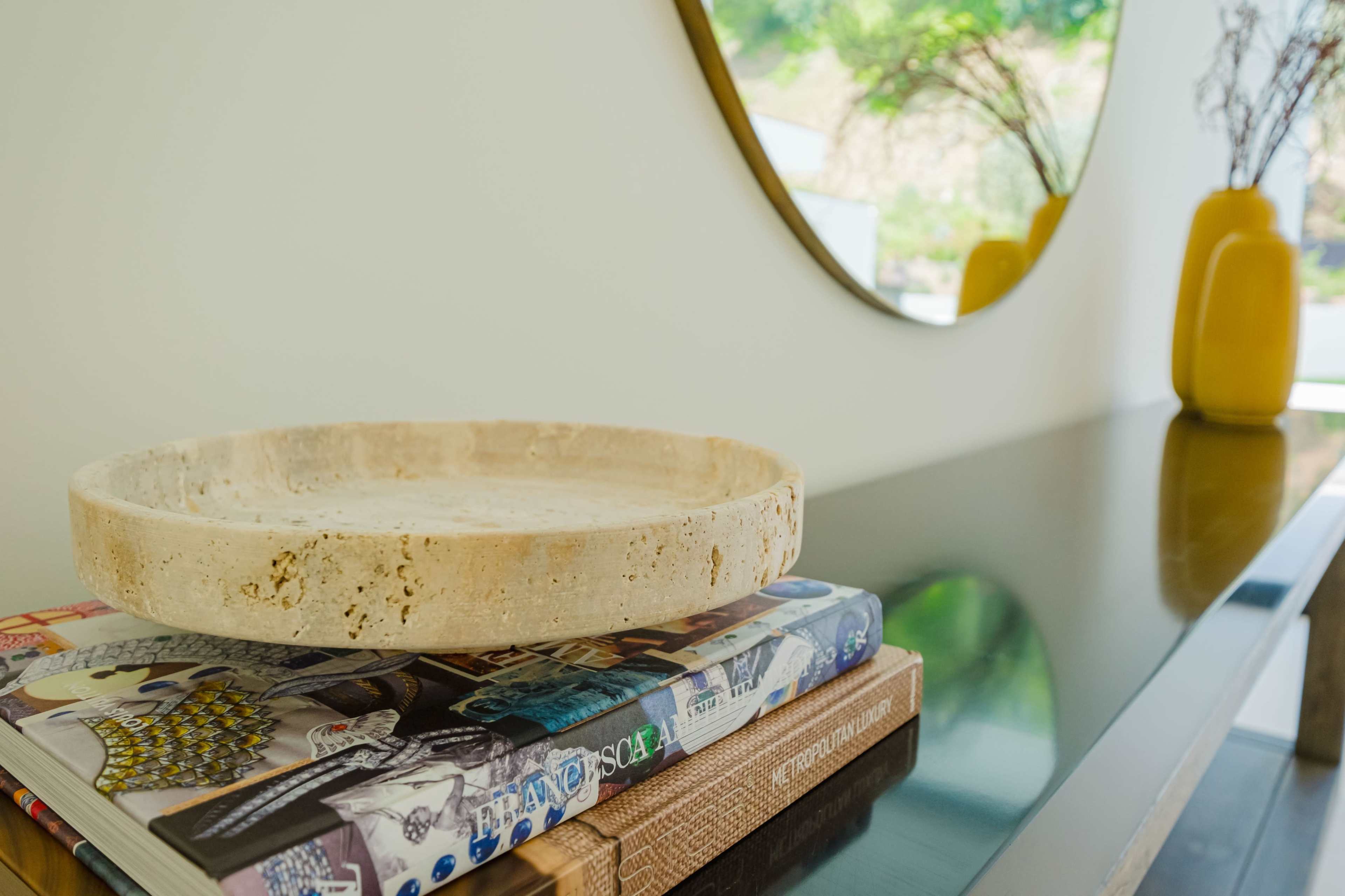 A round stone tray sits atop stacked magazines on a glossy table, with a large mirror reflecting a green landscape in the background.