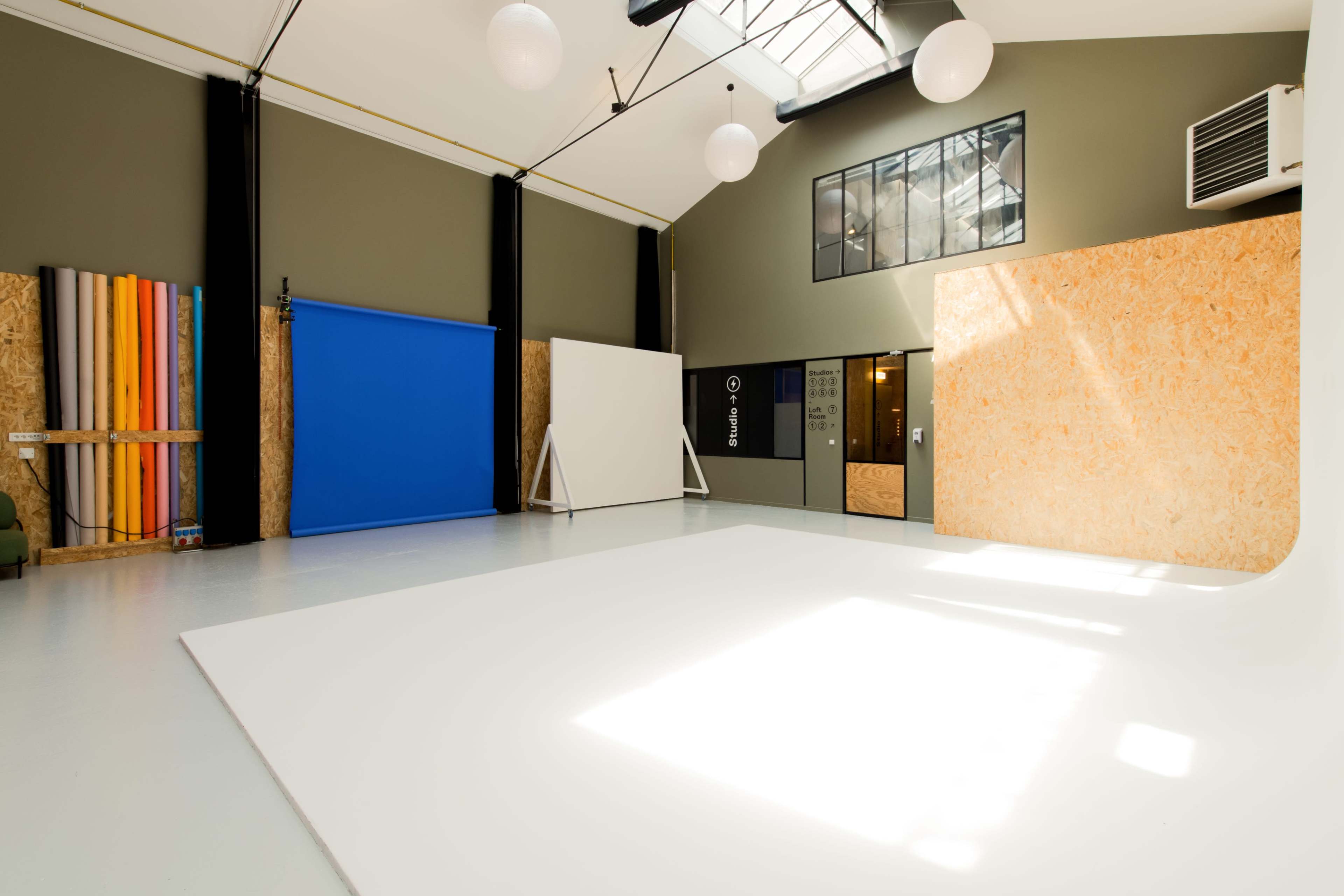 A spacious studio with white flooring, a blue backdrop, a large light-colored wall, and colorful vertical panels along with large windows and air conditioning.