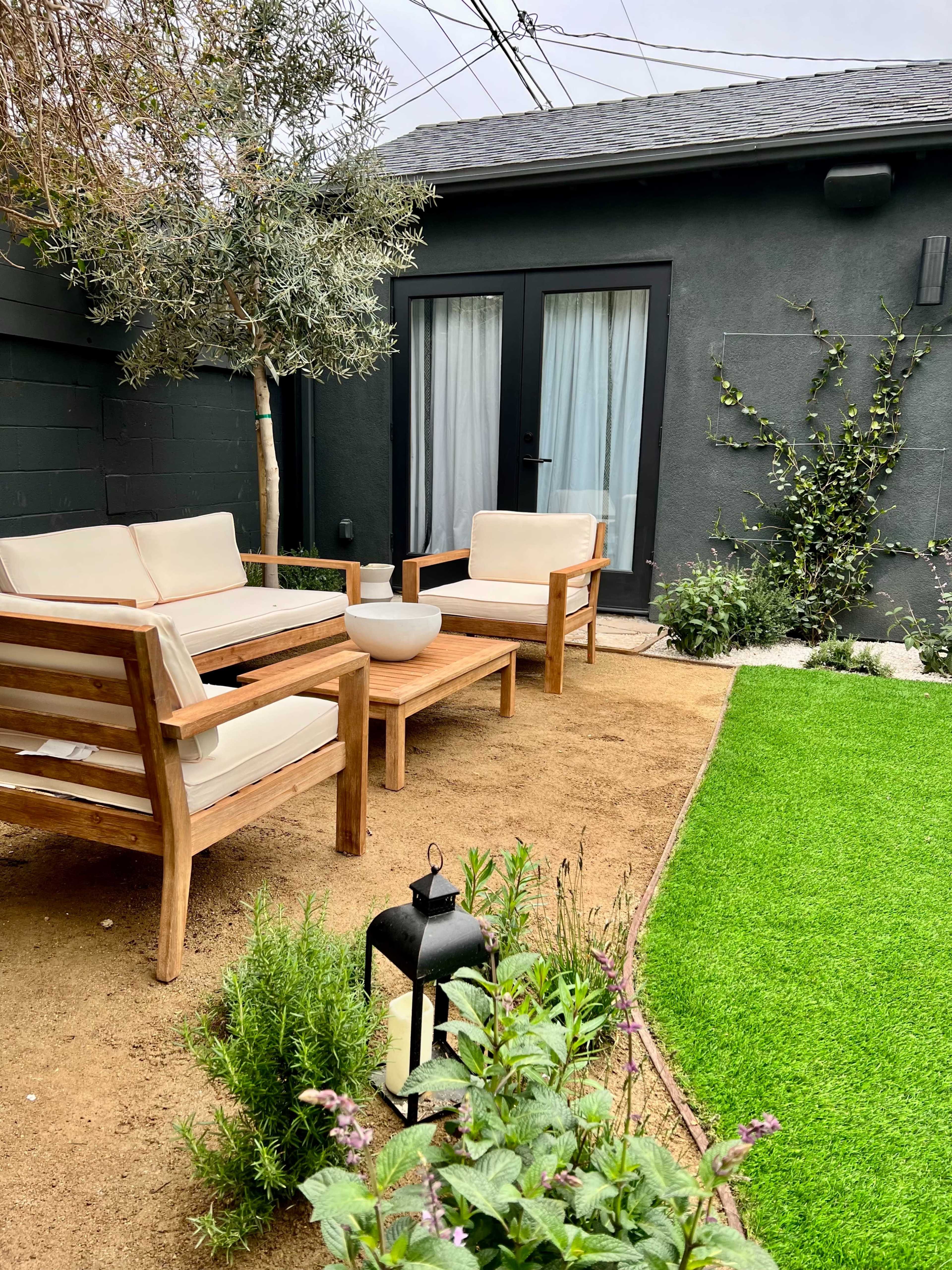 A small outdoor area features two wooden chairs with cushions, a round table, and a patch of artificial grass surrounded by plants and a lantern.