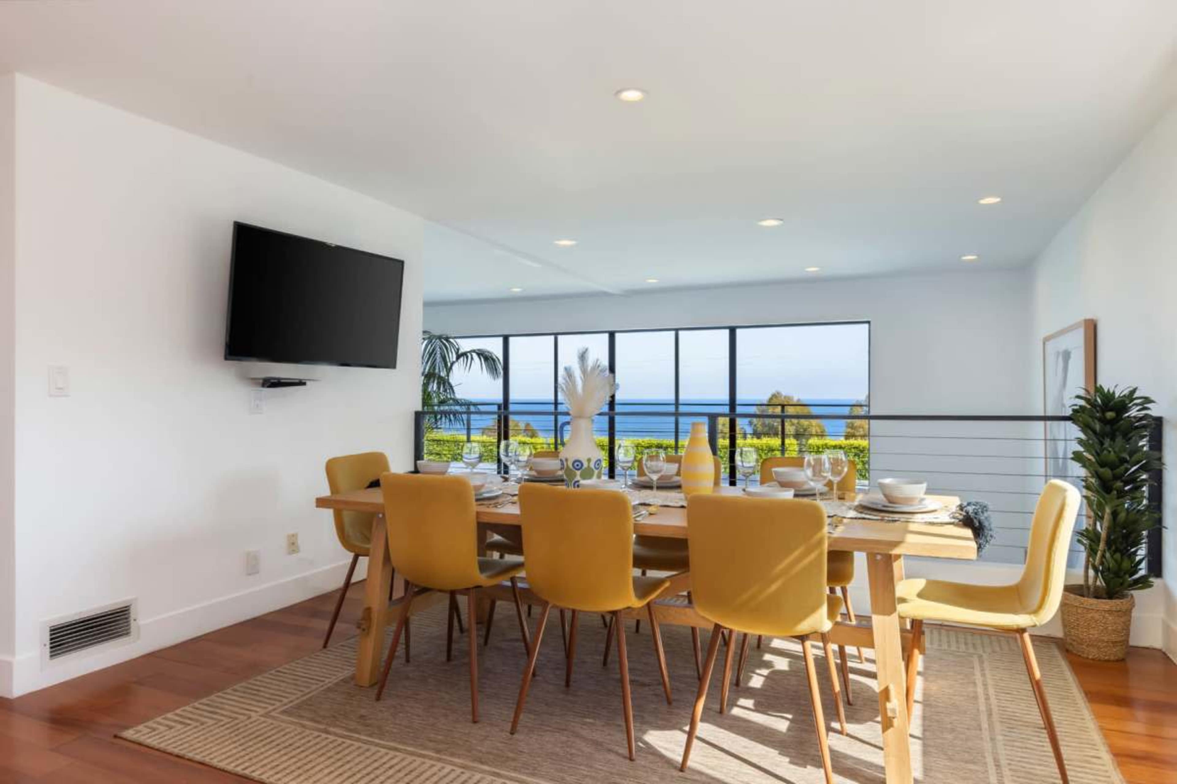 A modern dining room features a wooden table set for eight, with yellow chairs and a large window overlooking a water view.