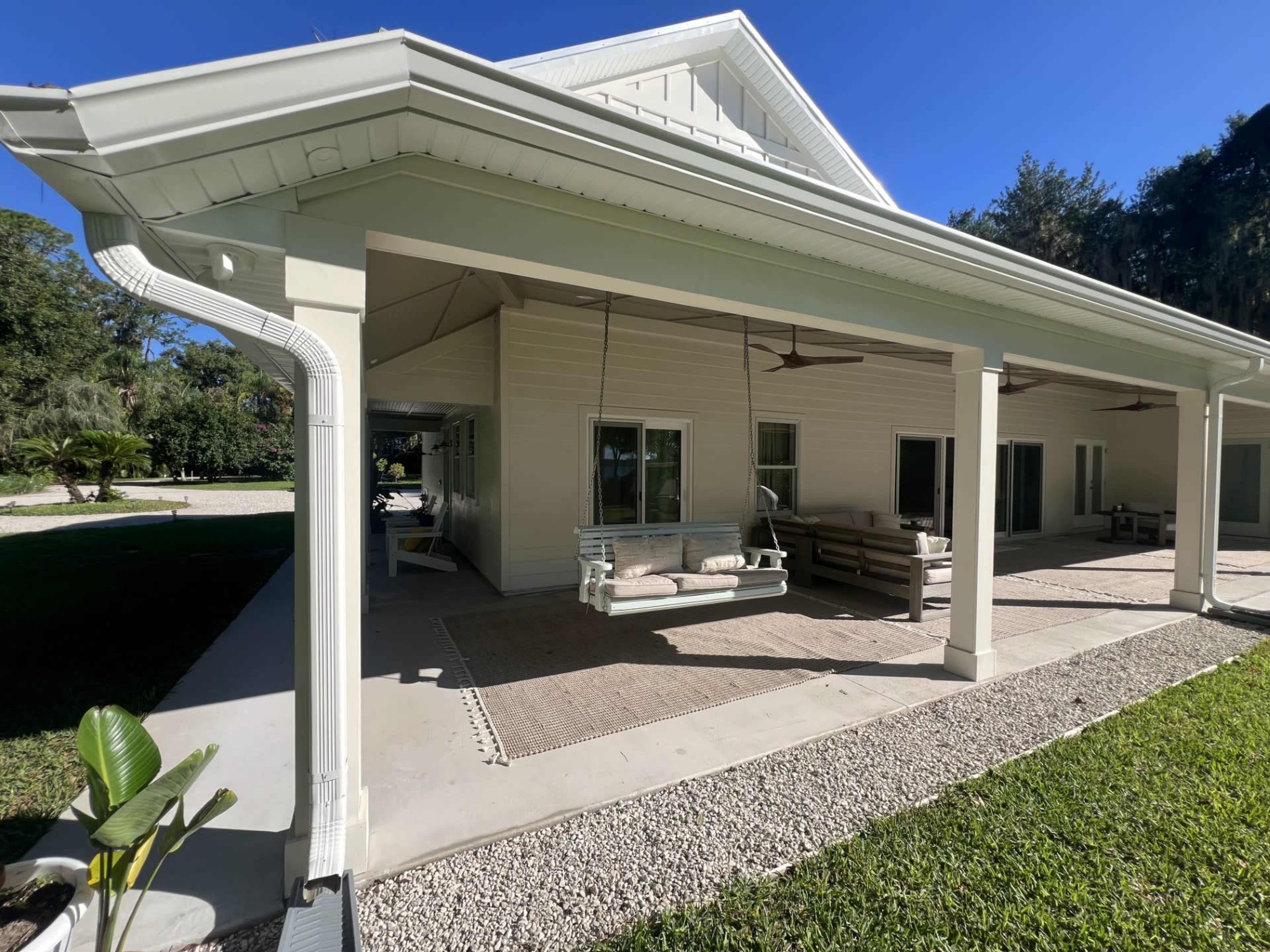 The image shows a spacious porch area of a house with a swing and seating arrangement, surrounded by green grass and trees.