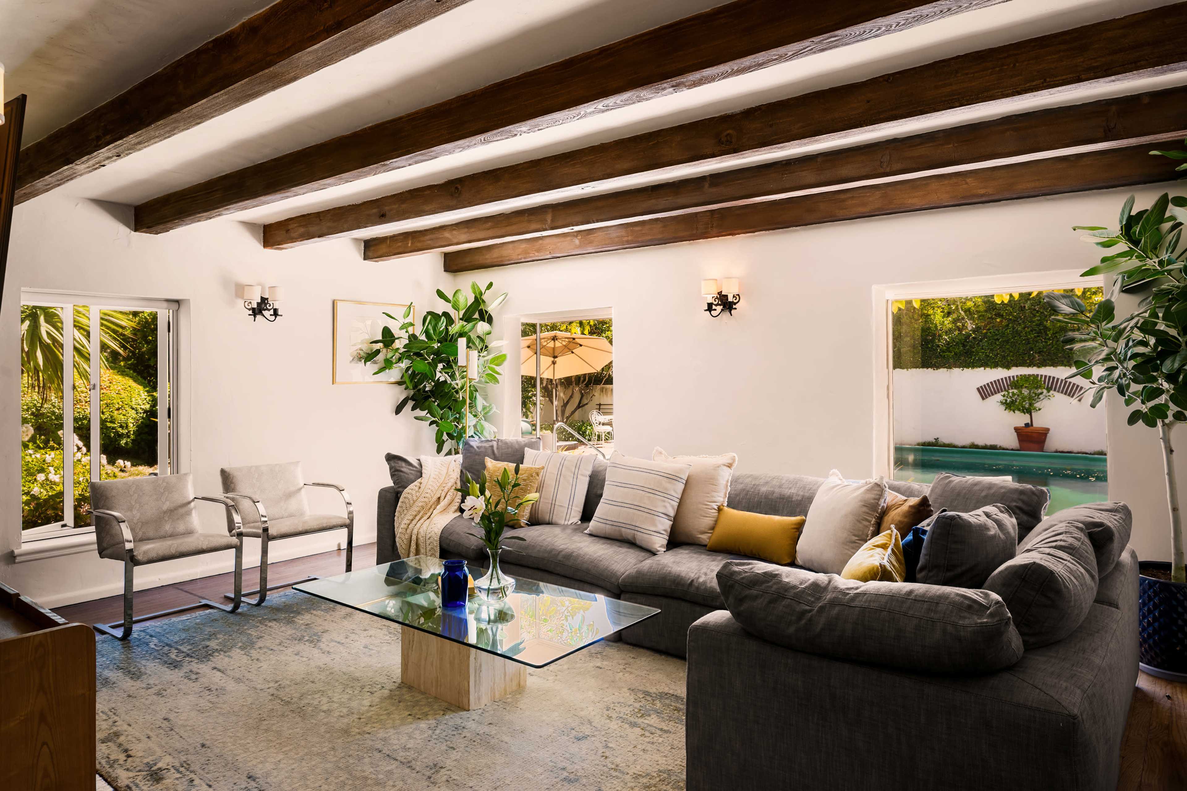 The living room features a spacious gray sofa, a glass coffee table, and large windows overlooking a pool area.