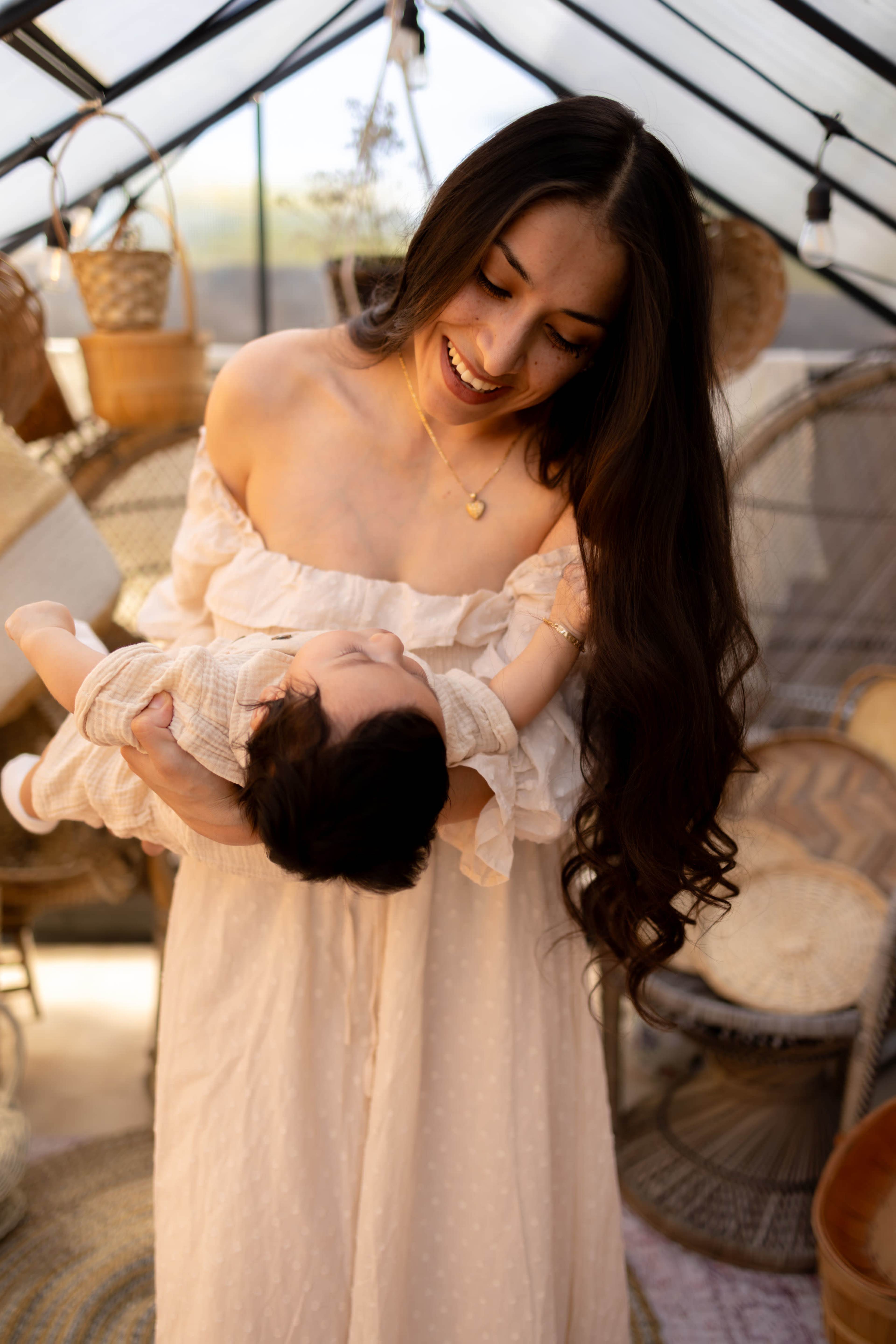 A woman in a light-colored dress holds a baby in a glass greenhouse filled with plants and decorative baskets.