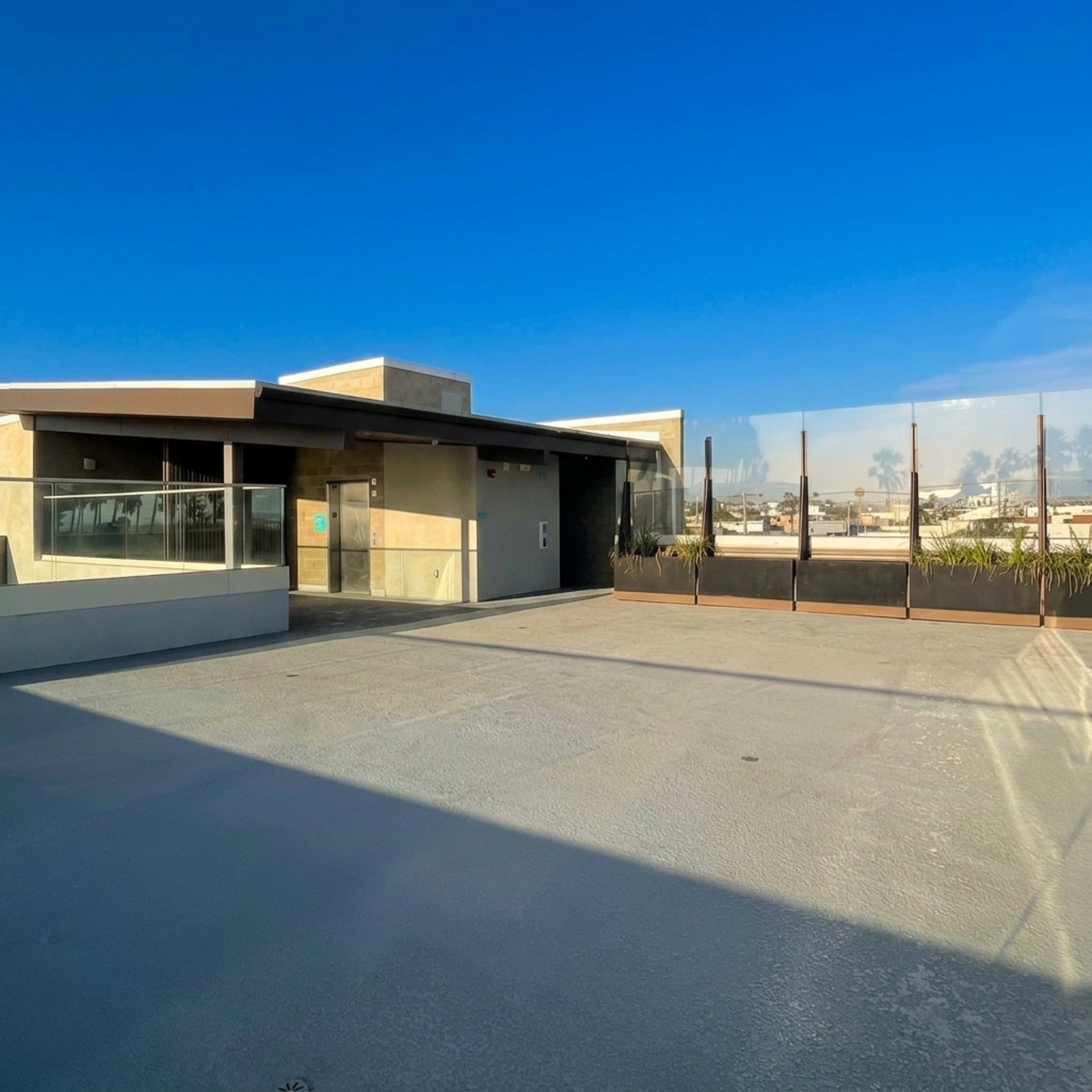 The image shows a modern building with large glass windows and a paved outdoor area under a clear blue sky.