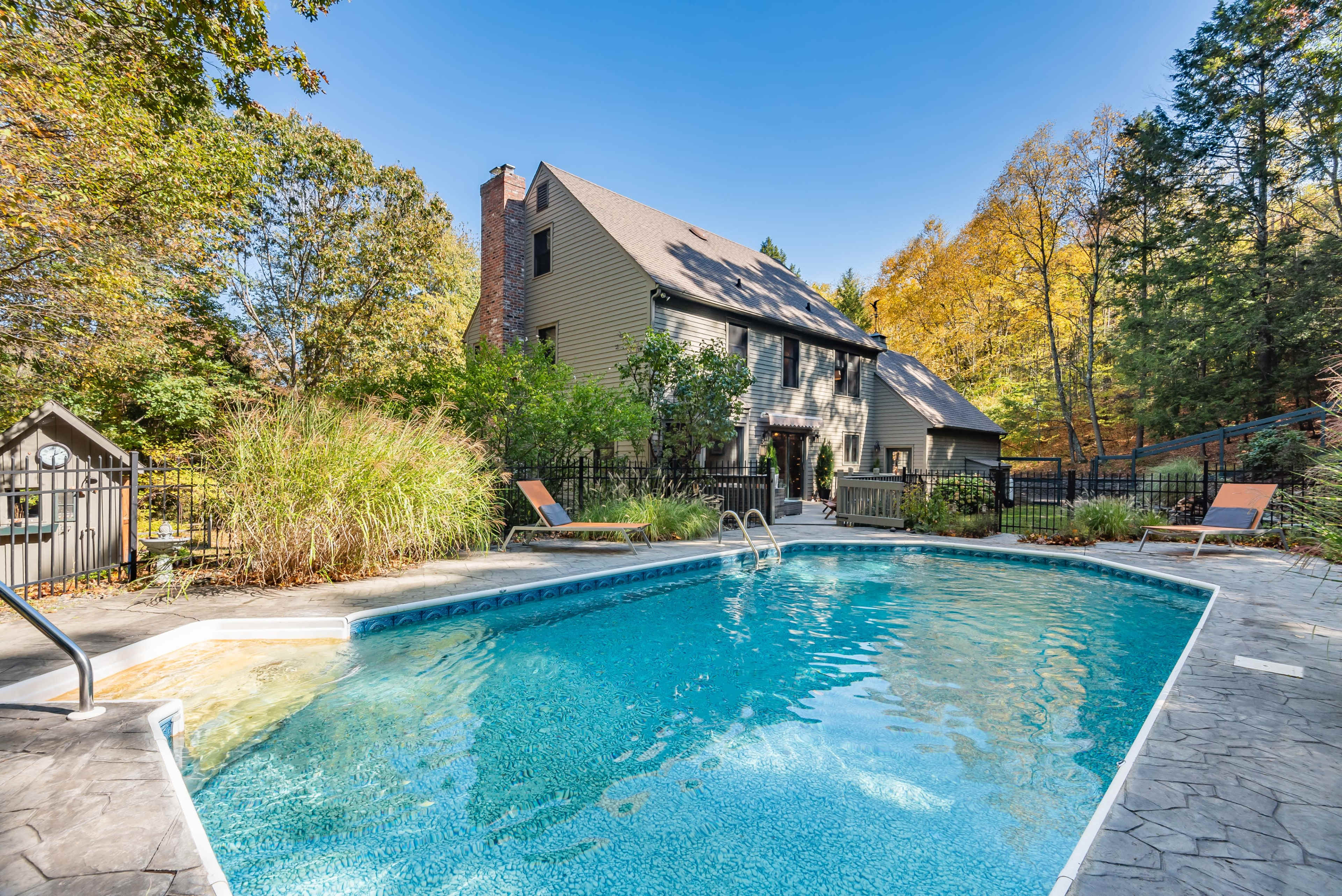 A swimming pool with a stone deck is situated next to a large house surrounded by trees in autumn foliage.