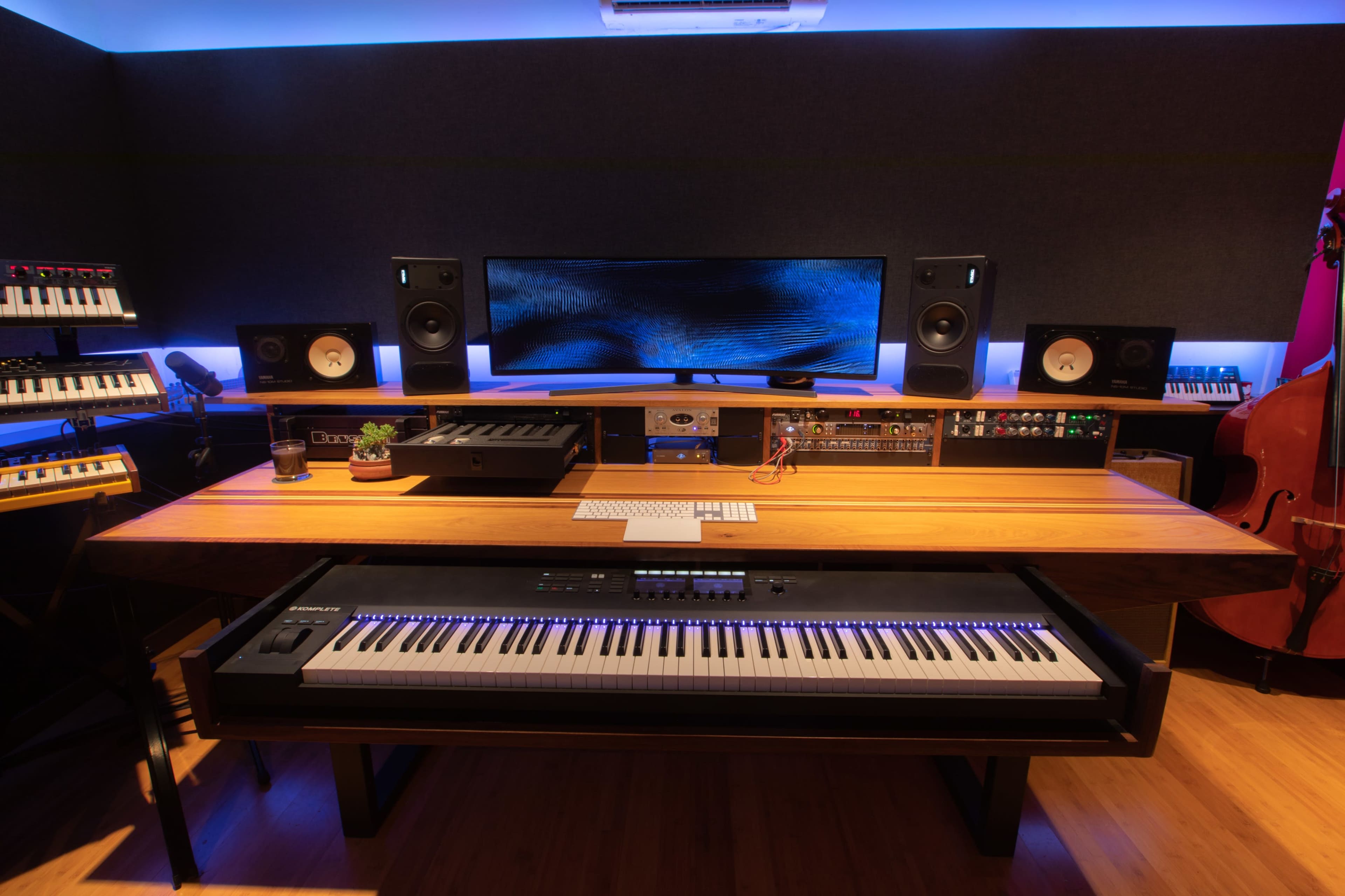 A modern music studio featuring a grand piano, audio equipment, a large monitor, and various keyboards arranged around a wooden tabletop.