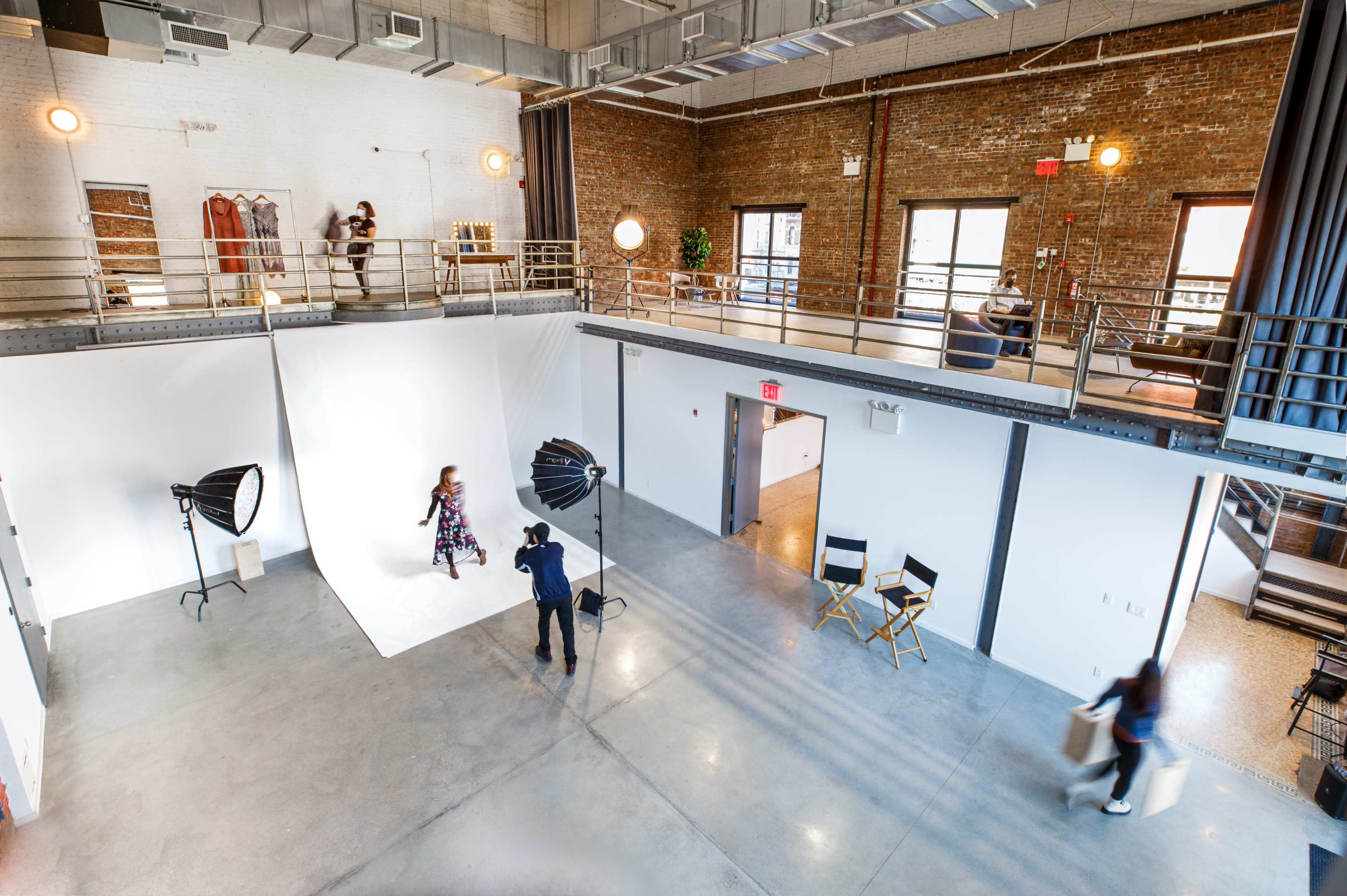 A spacious studio features a model posing on a white backdrop while a photographer captures the shot, with additional areas for changing clothes and seating visible in the background.