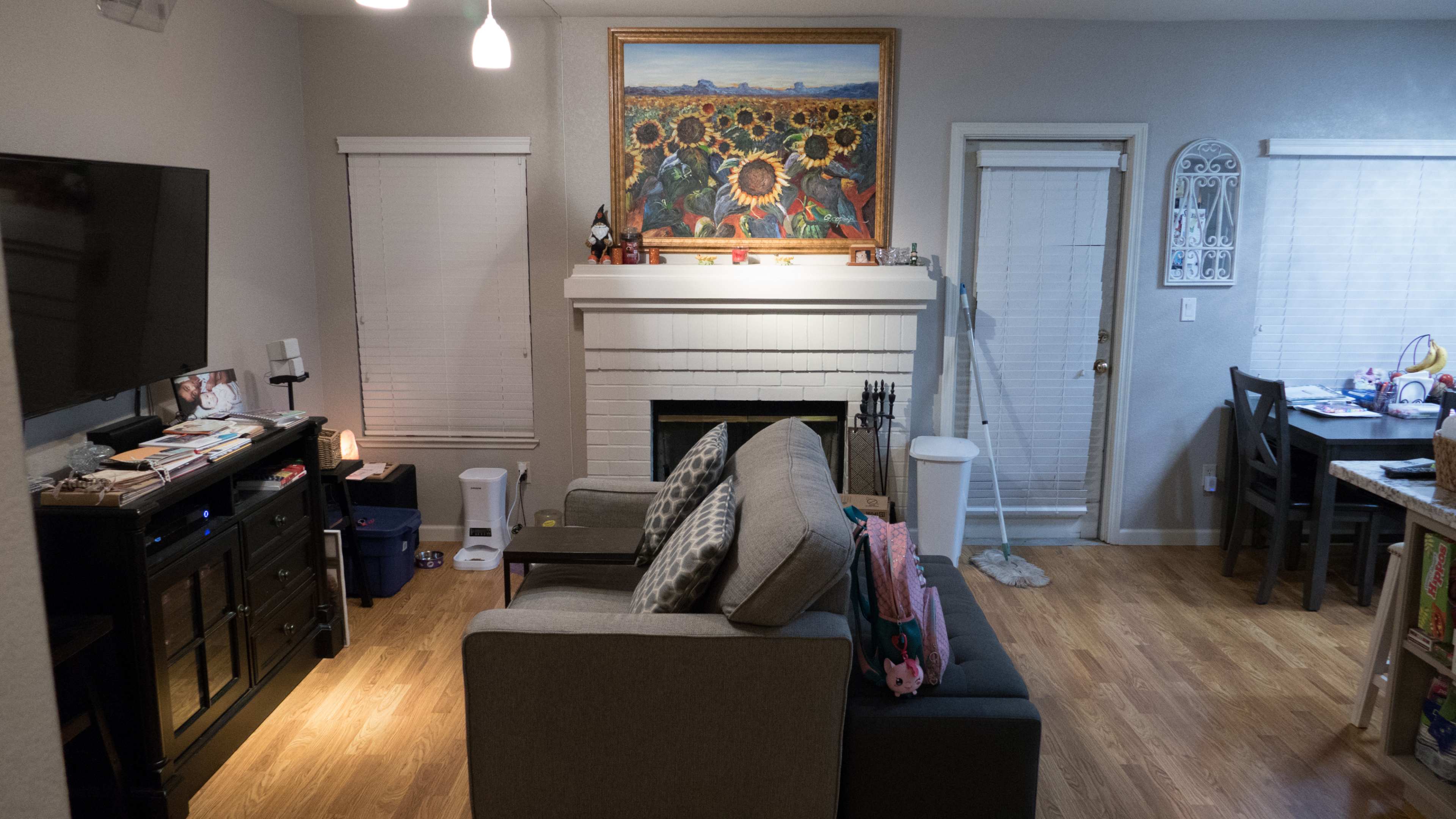 A cozy living room features a gray sofa, a television on a black cabinet, a fireplace adorned with a colorful sunflower painting, and a dining area with a table in the background.