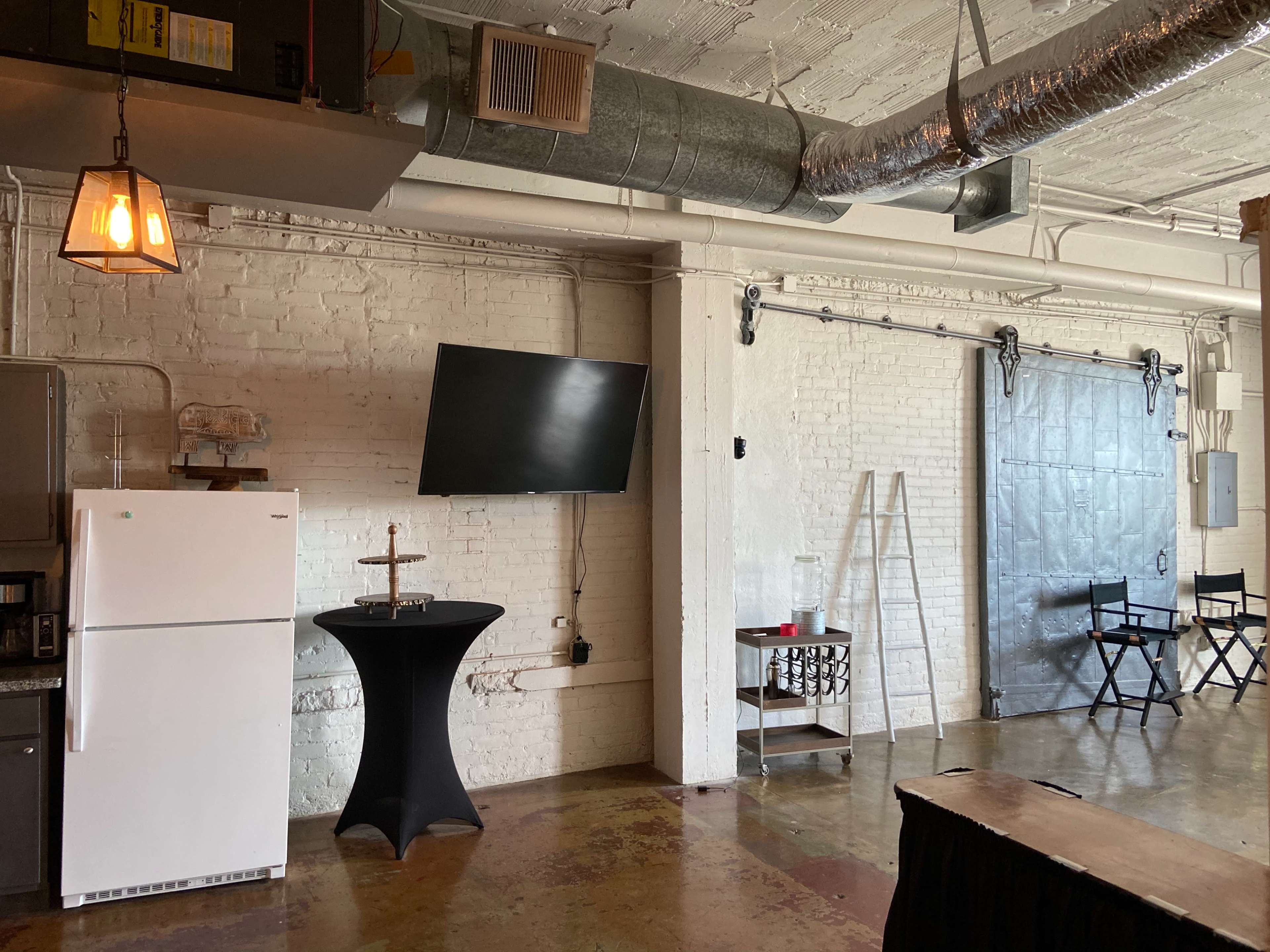 An industrial-style room with a white brick wall, a television mounted on the wall, a refrigerator, a black table, and a sliding door.