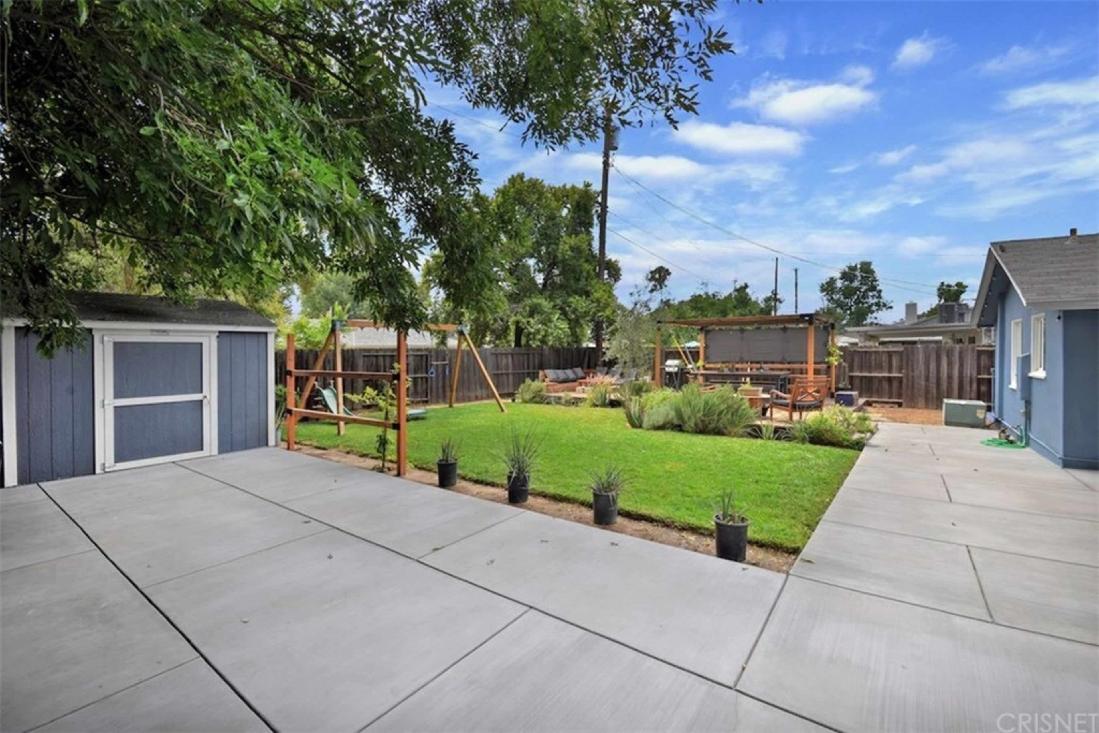 The image shows a spacious backyard with a grassy area, playset, garden, and two concrete patios.