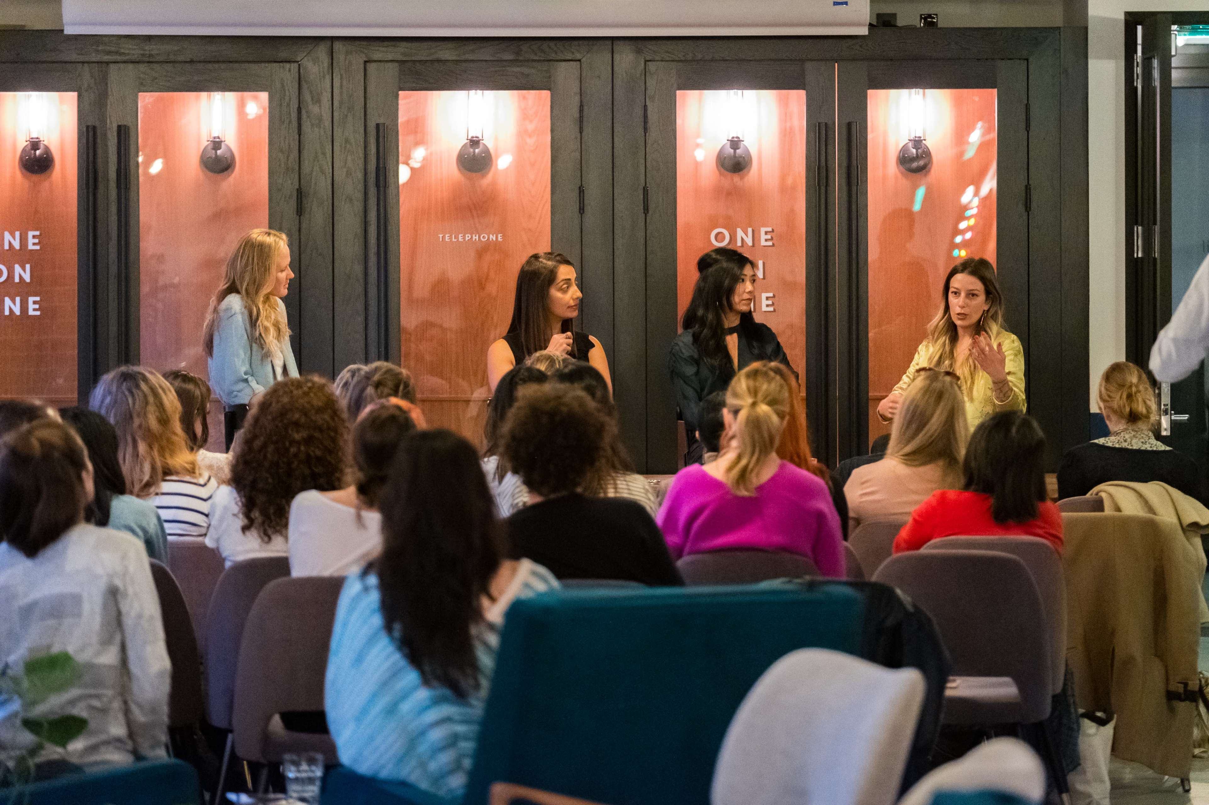 A panel of four women is engaged in discussion in front of an audience seated in a modern room with wooden accents.
