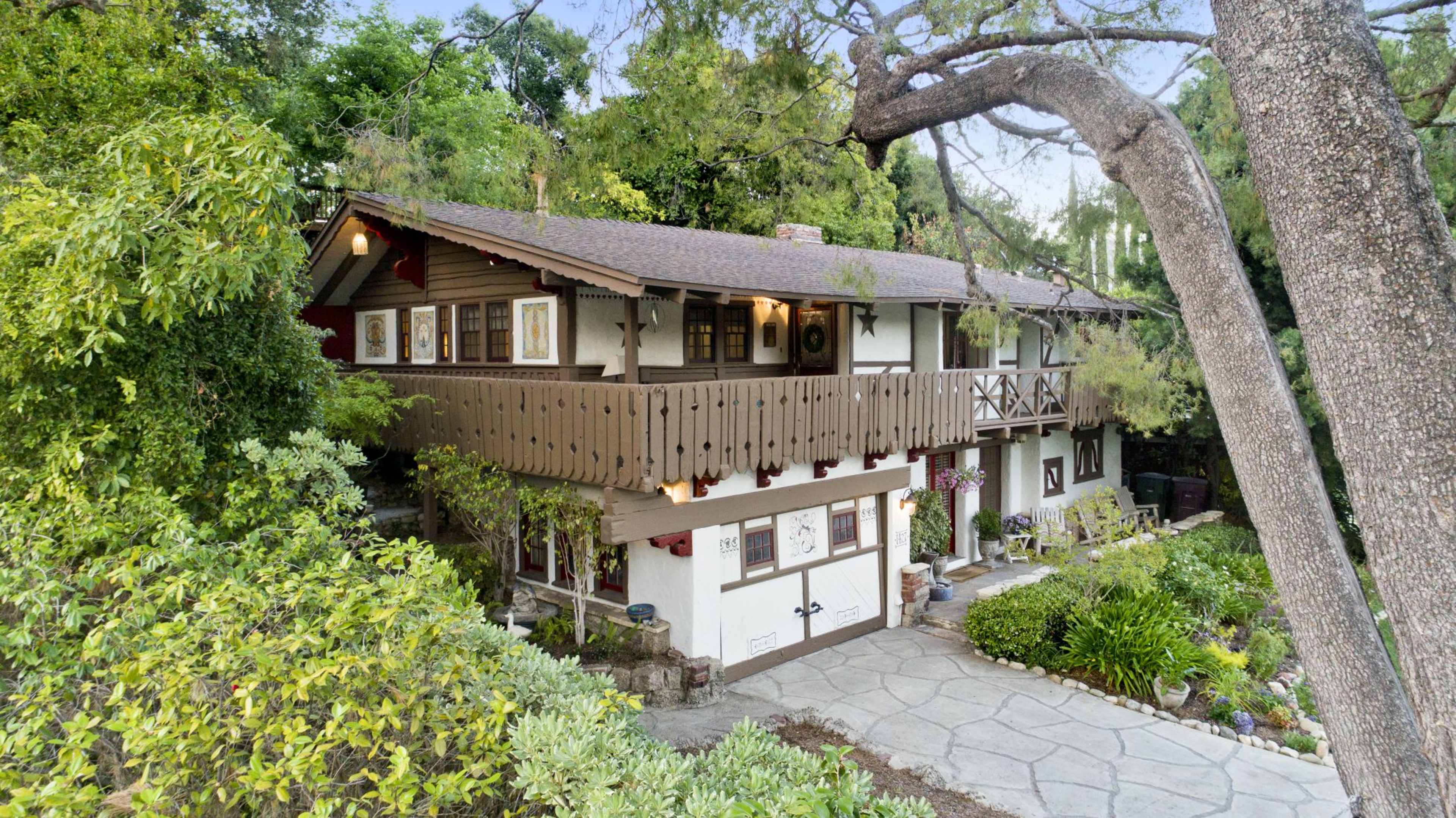 A two-story house with a balcony and wooden accents is surrounded by lush greenery and a stone pathway.