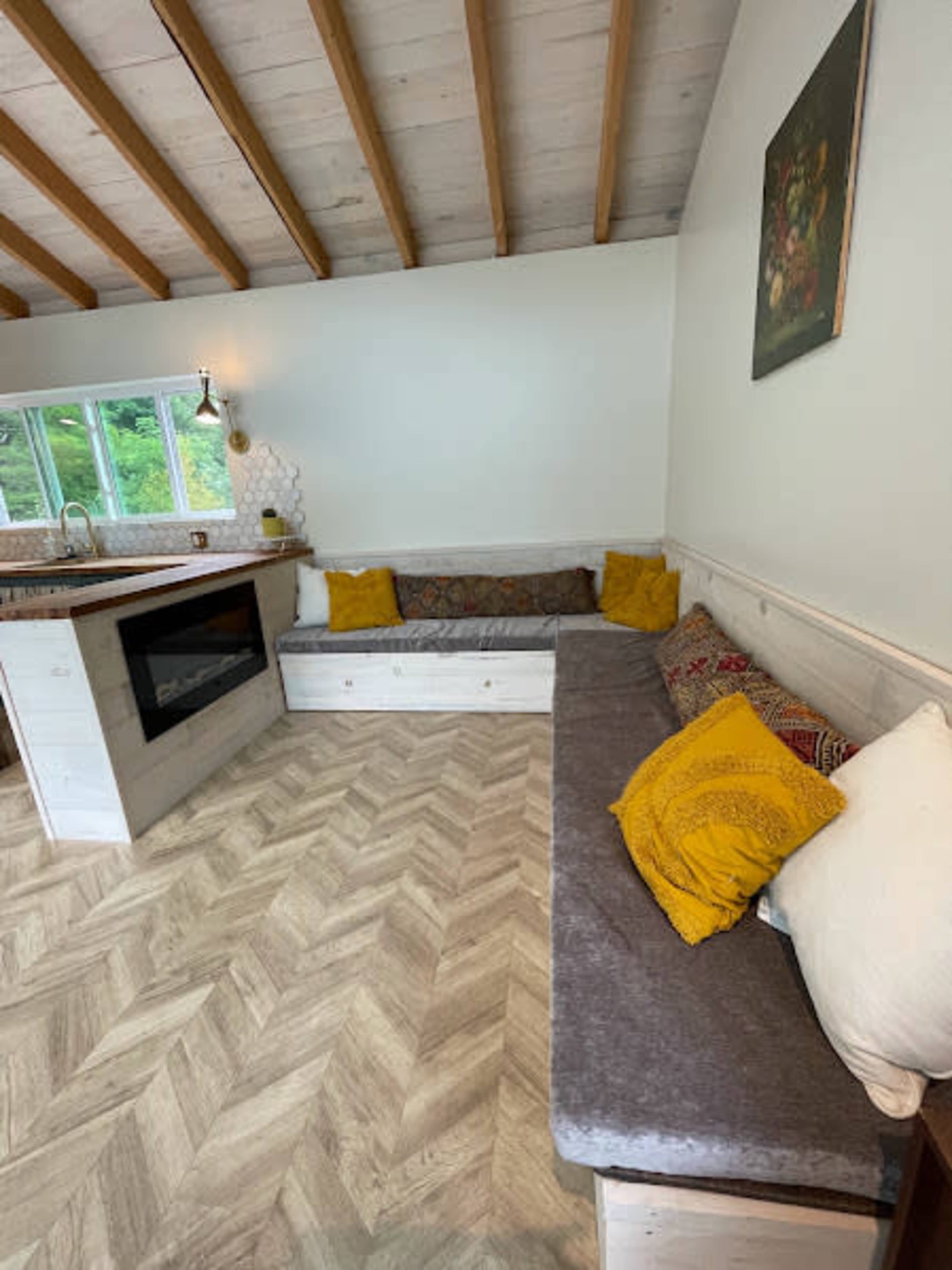 The image shows a cozy living area with a L-shaped seating arrangement, featuring a mix of cushions and a small fireplace beside a kitchen space.