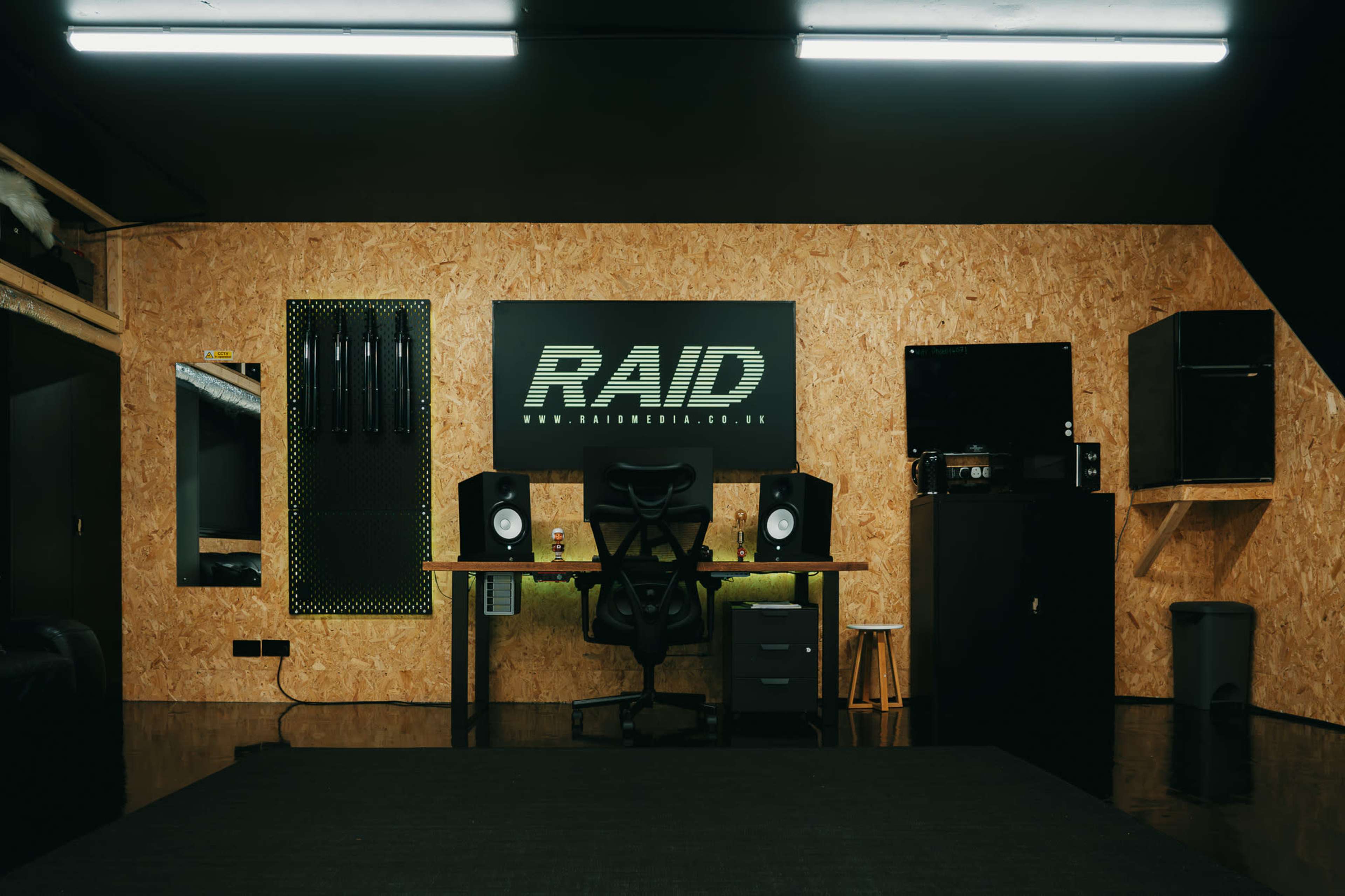 A modern workspace features a desk with audio monitors, a black chair, and a large RAID logo on an oriented strand board wall.
