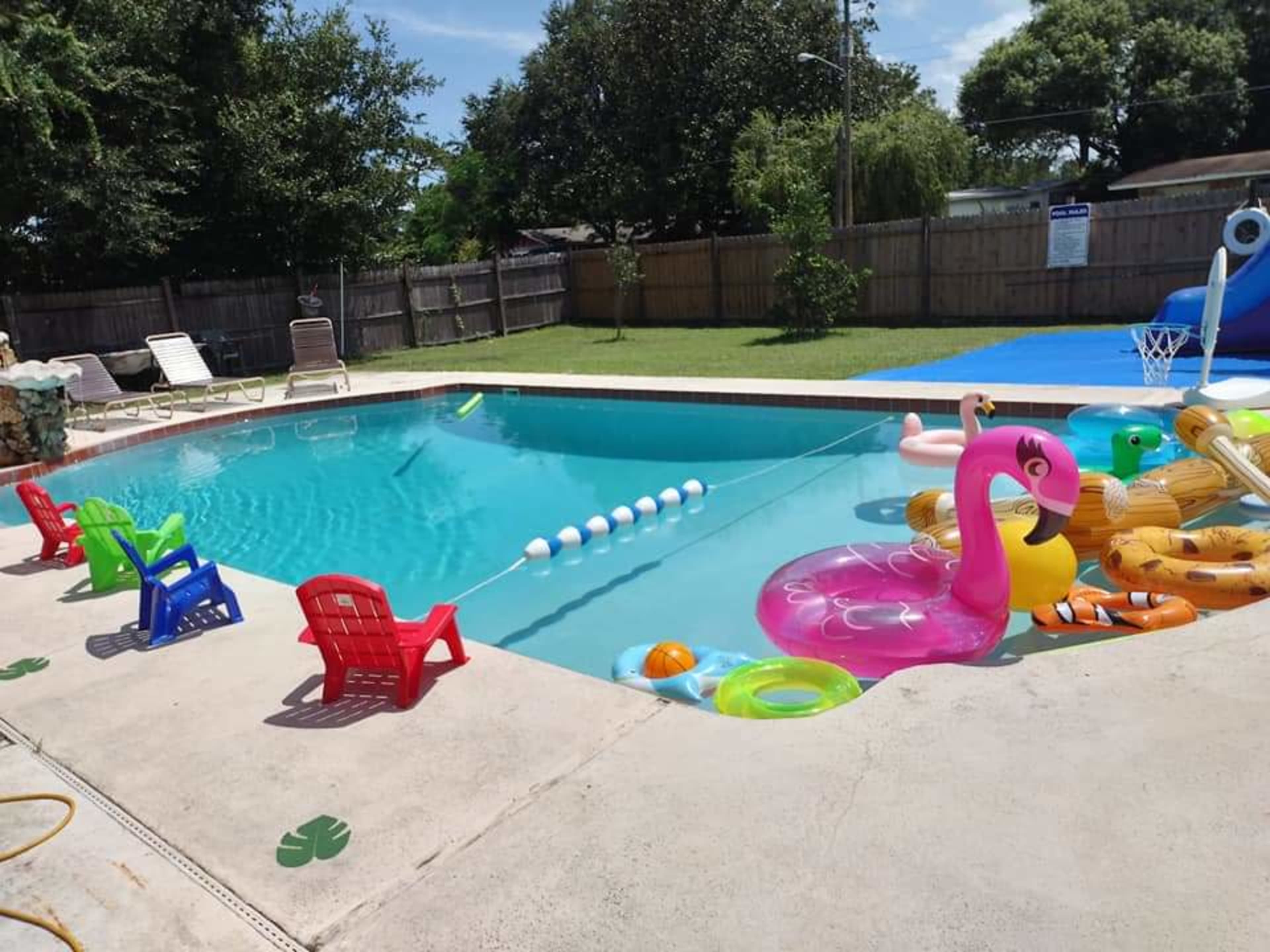 A backyard swimming pool is surrounded by colorful inflatable floats and small plastic chairs on the concrete deck.