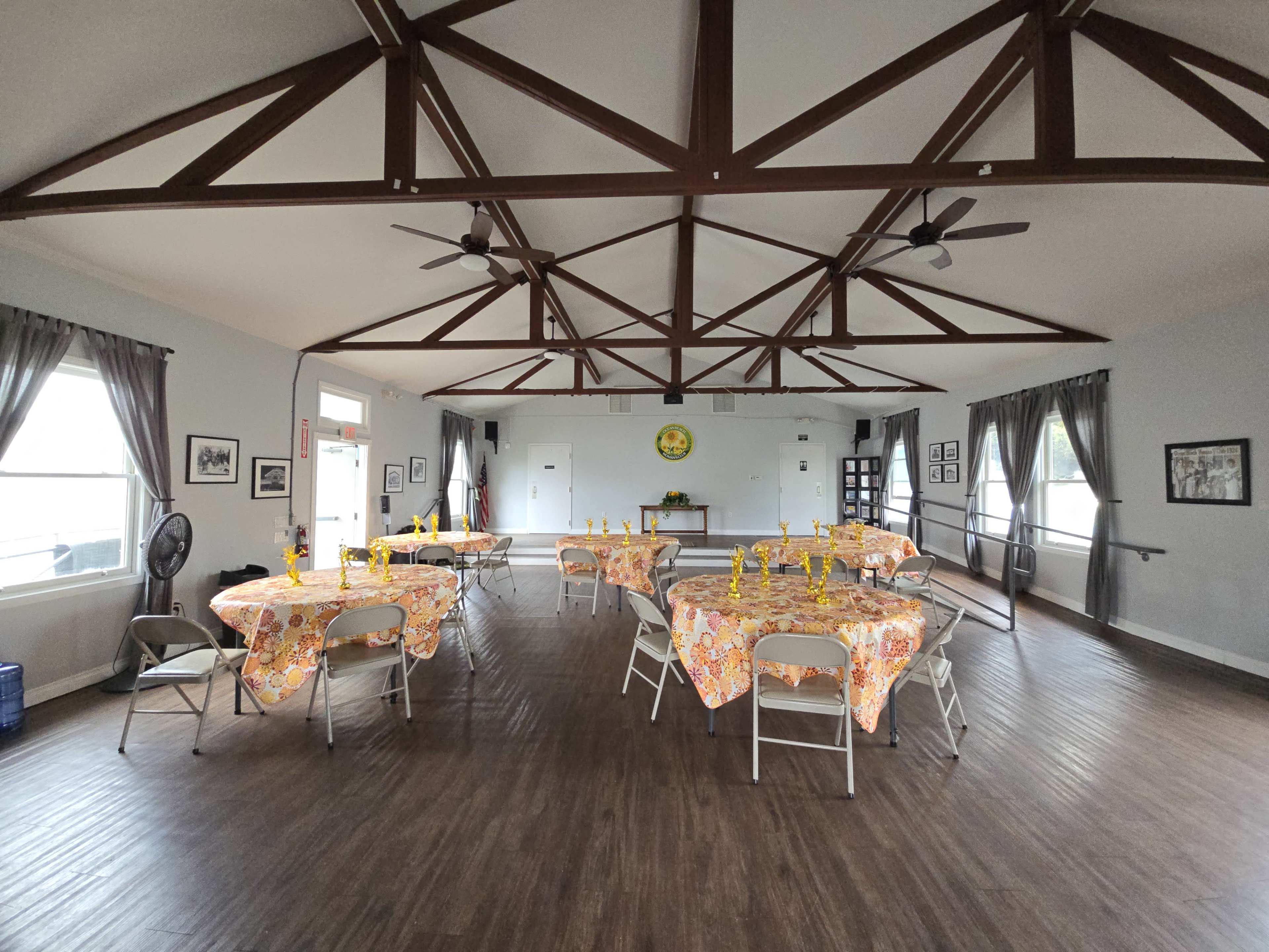 The image shows a spacious room with several tables covered in floral tablecloths, arranged for a gathering, beneath a wooden beam ceiling.