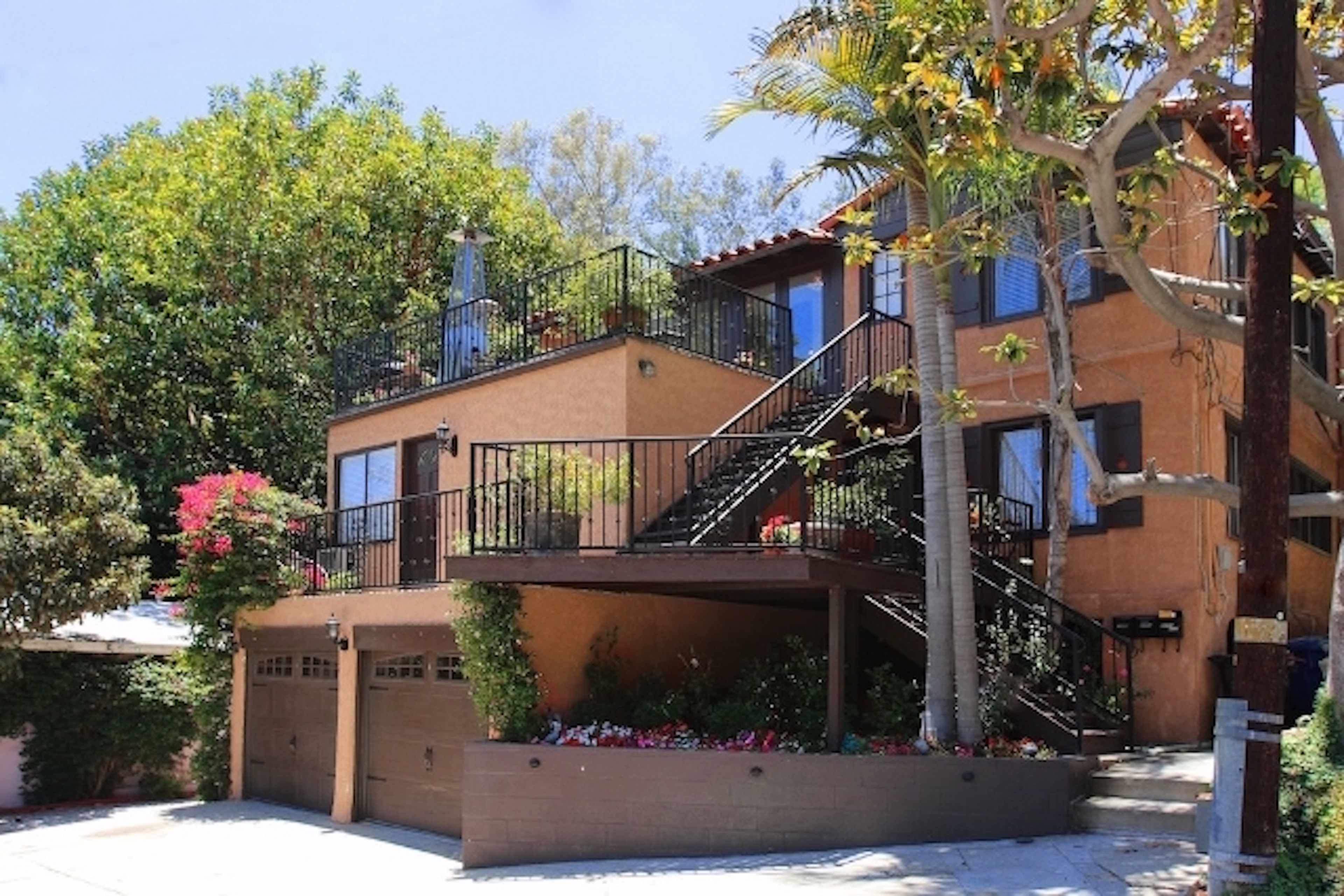A two-story residential building with a balcony, surrounded by greenery and colorful flowers.